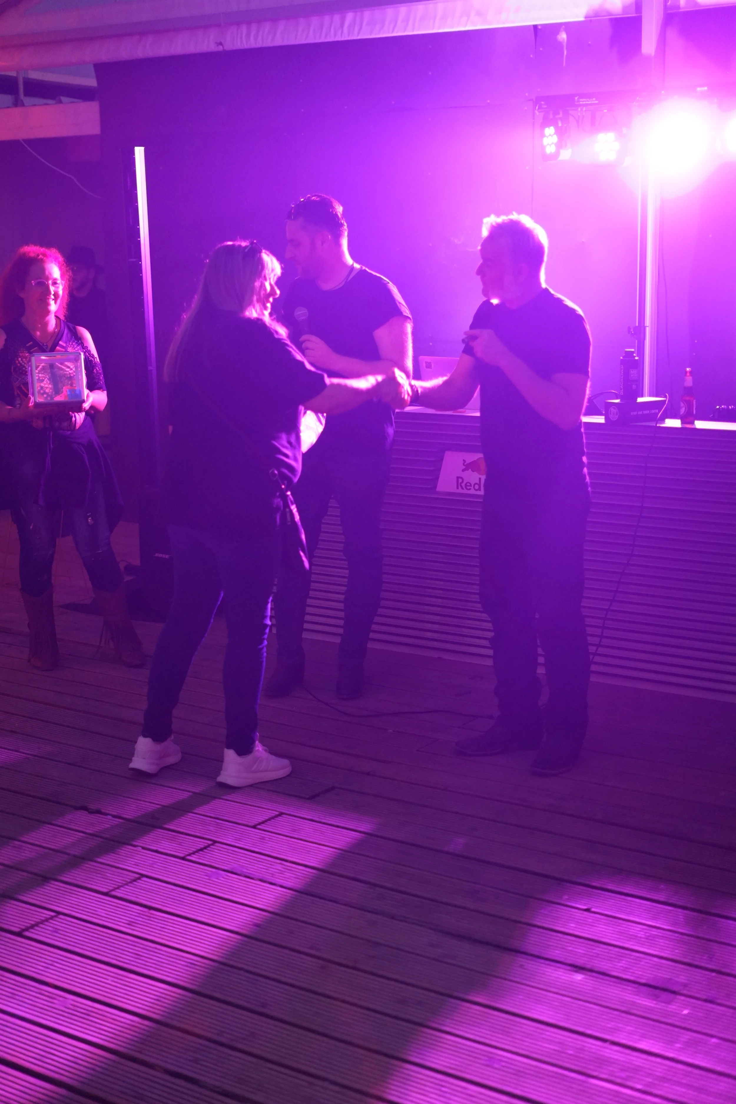 Four people on a dance floor under purple and pink lighting. Two women are dancing and two men are nearby, one holding a microphone. One woman is holding a box with a glass window, and the background includes DJ equipment and a 