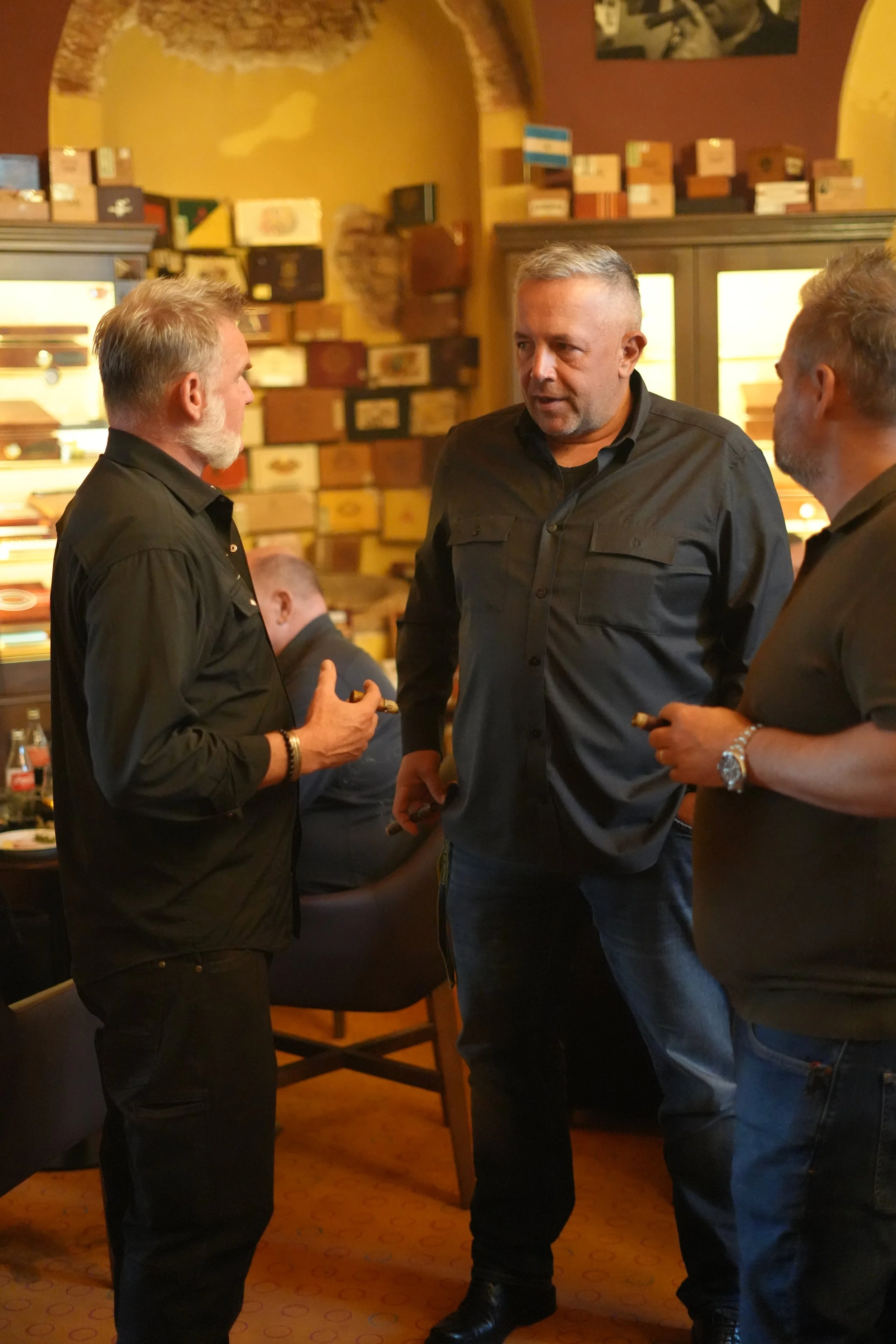 Three men are standing and talking in a dimly lit room with a yellow ceiling and walls decorated with cigars and cigar boxes. One man is holding a cigar and is speaking to a man with short gray hair and a beard. The third man is partially visible on 