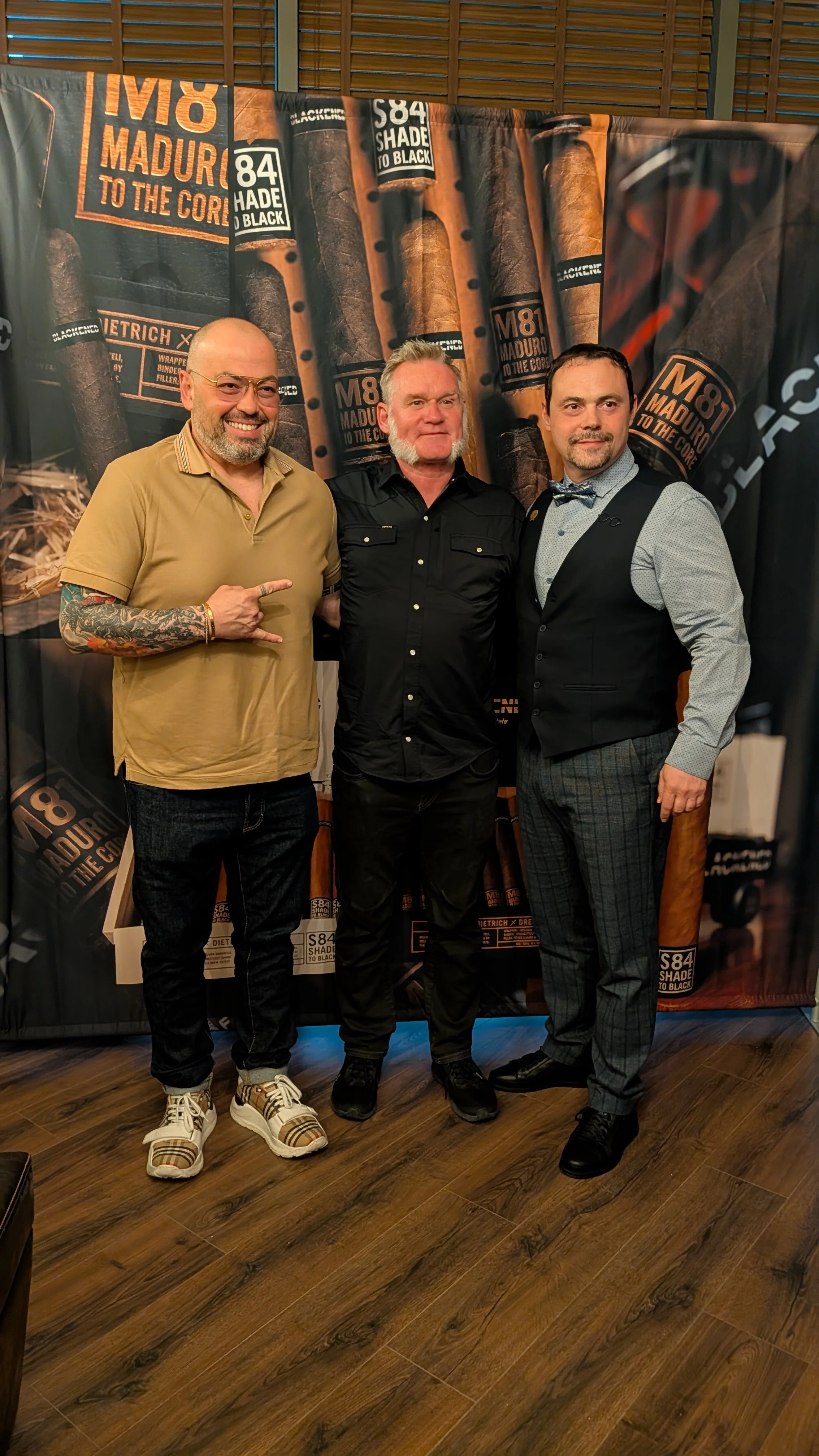 Three men standing in front of a backdrop with cigar images and text, smiling at the camera.