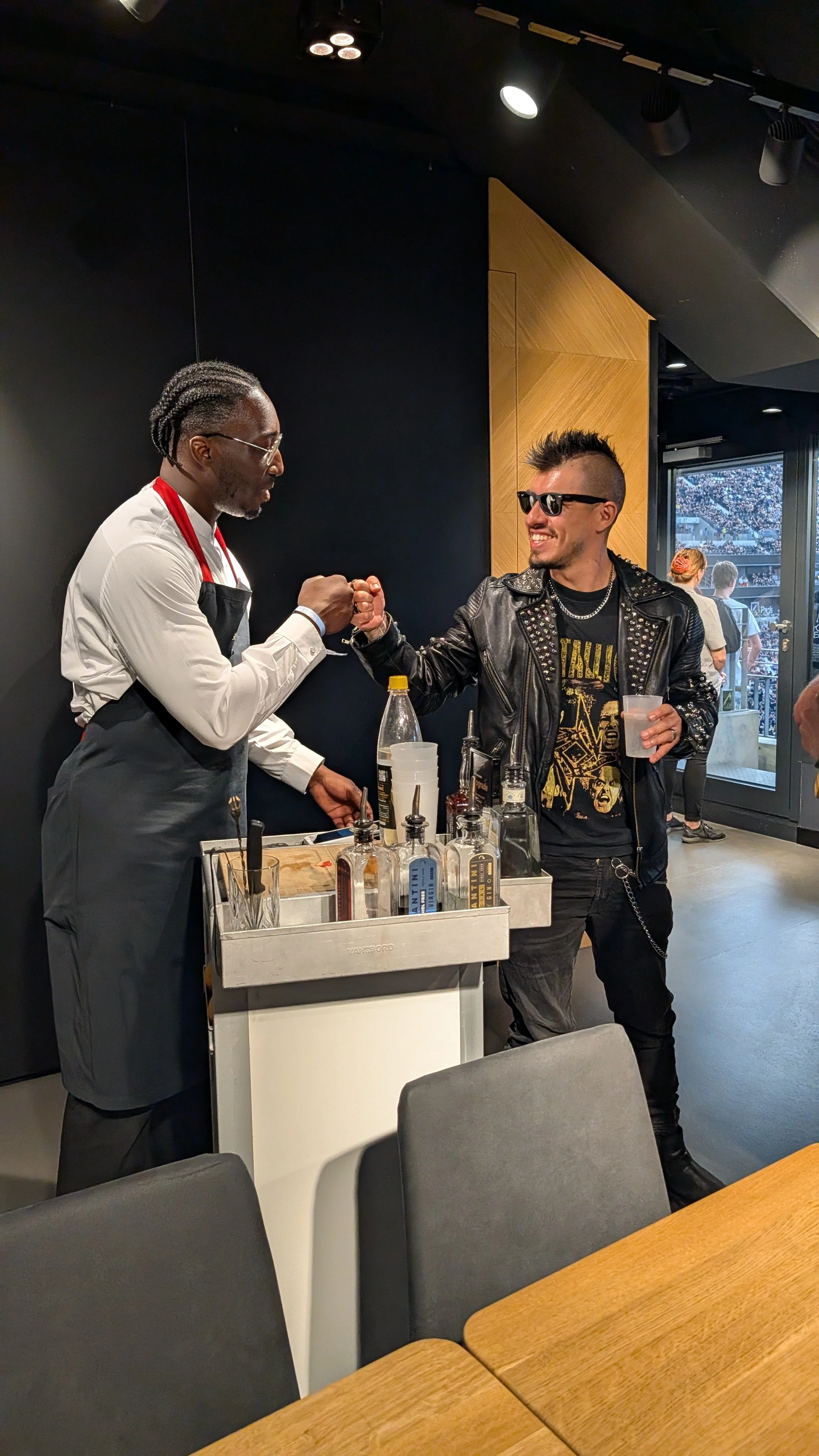 A bartender and a customer fist bump inside a modern bar or restaurant. The bartender is wearing a white shirt, gray apron, and glasses, while the customer has sunglasses, a leather jacket with studs, and is holding a drink. There are bottles and bar