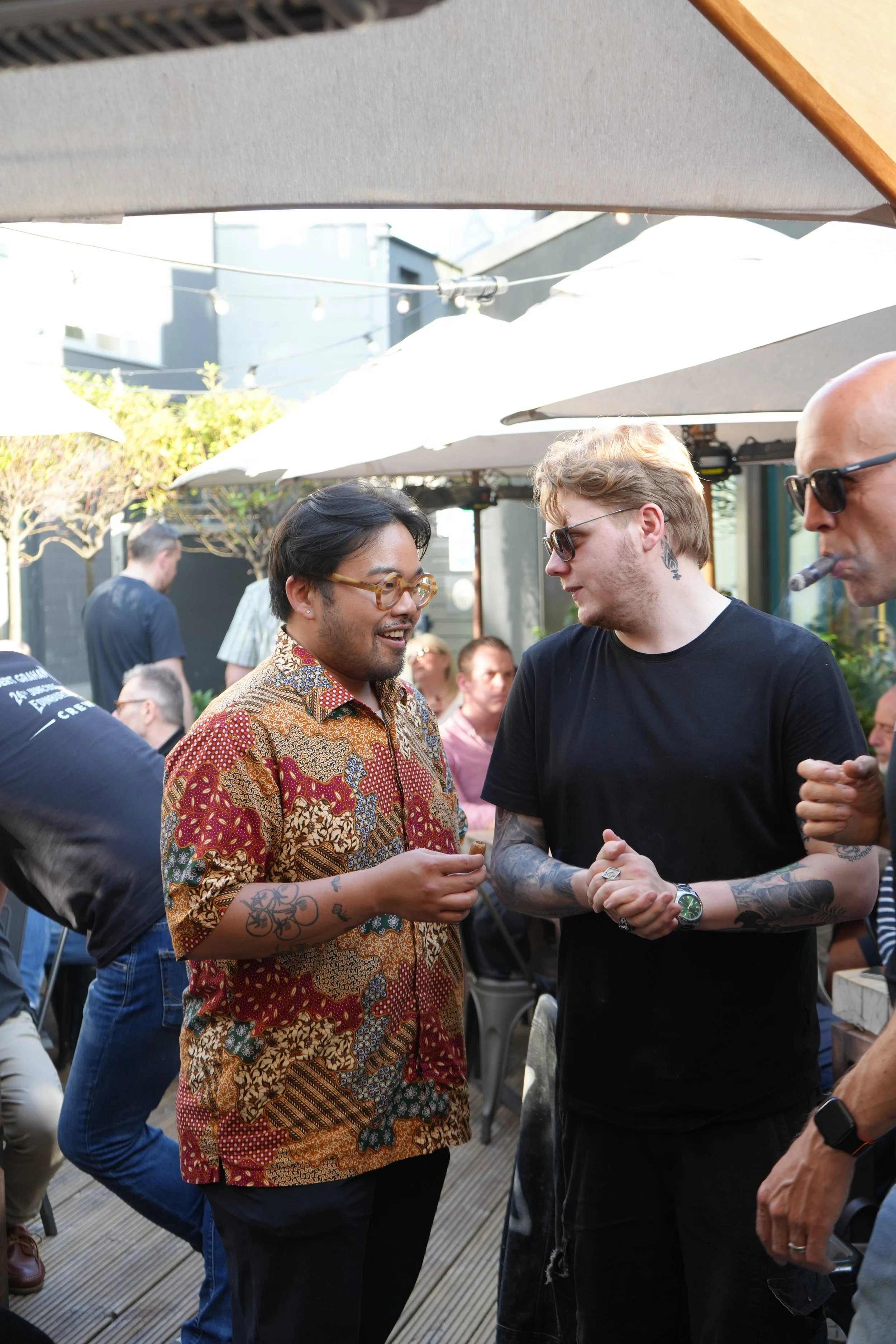 Two men, one wearing a colorful patterned shirt and glasses, and the other wearing a black shirt, sunglasses, and tattoos, shake hands at an outdoor gathering with umbrellas and other people in the background.