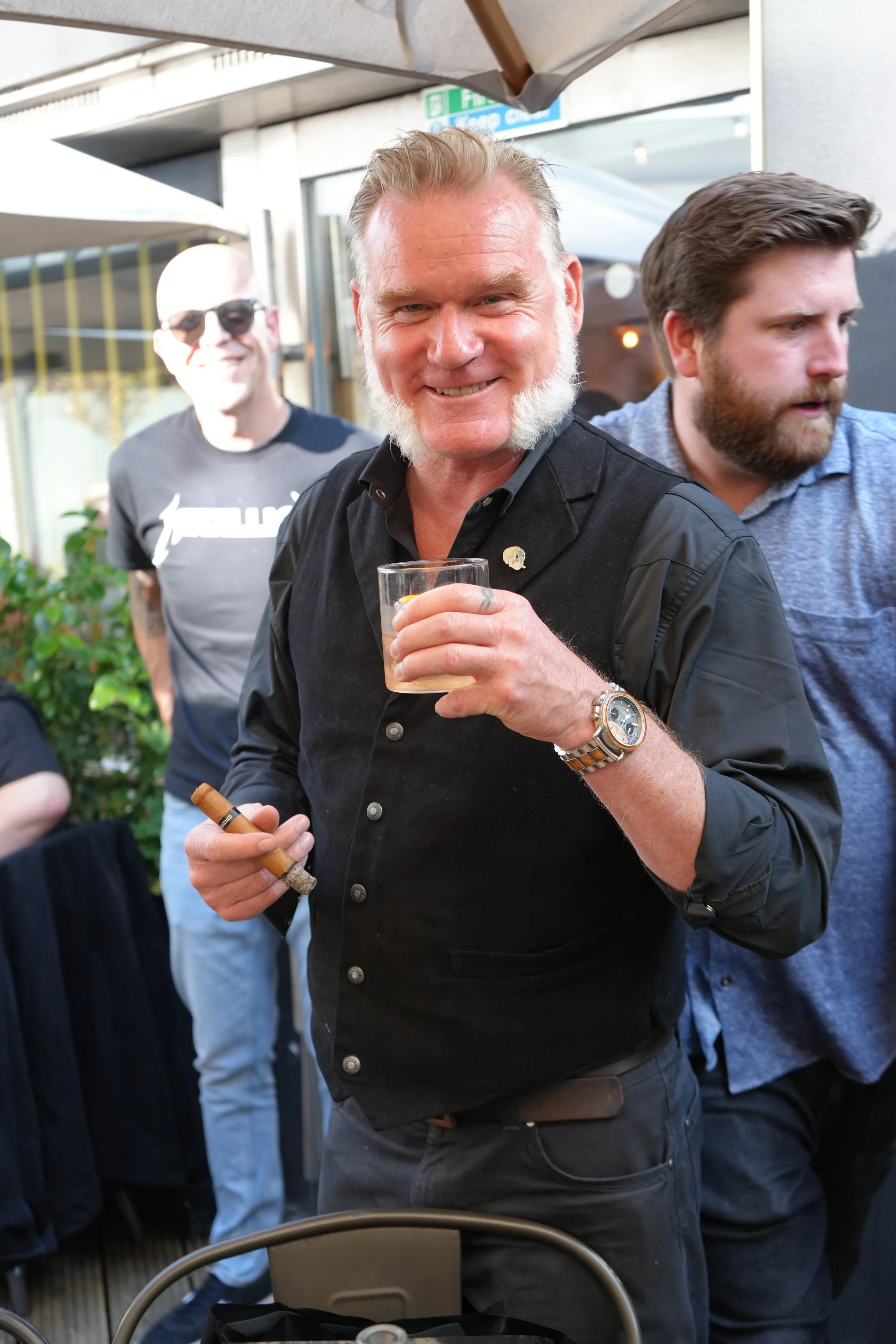 A group of men socializing outdoors, with one man in the foreground smiling, holding a cigar and a drink, wearing a black shirt, a wristwatch, and a small pin on his shirt. Others are in the background, one wearing sunglasses and a black t-shirt, the