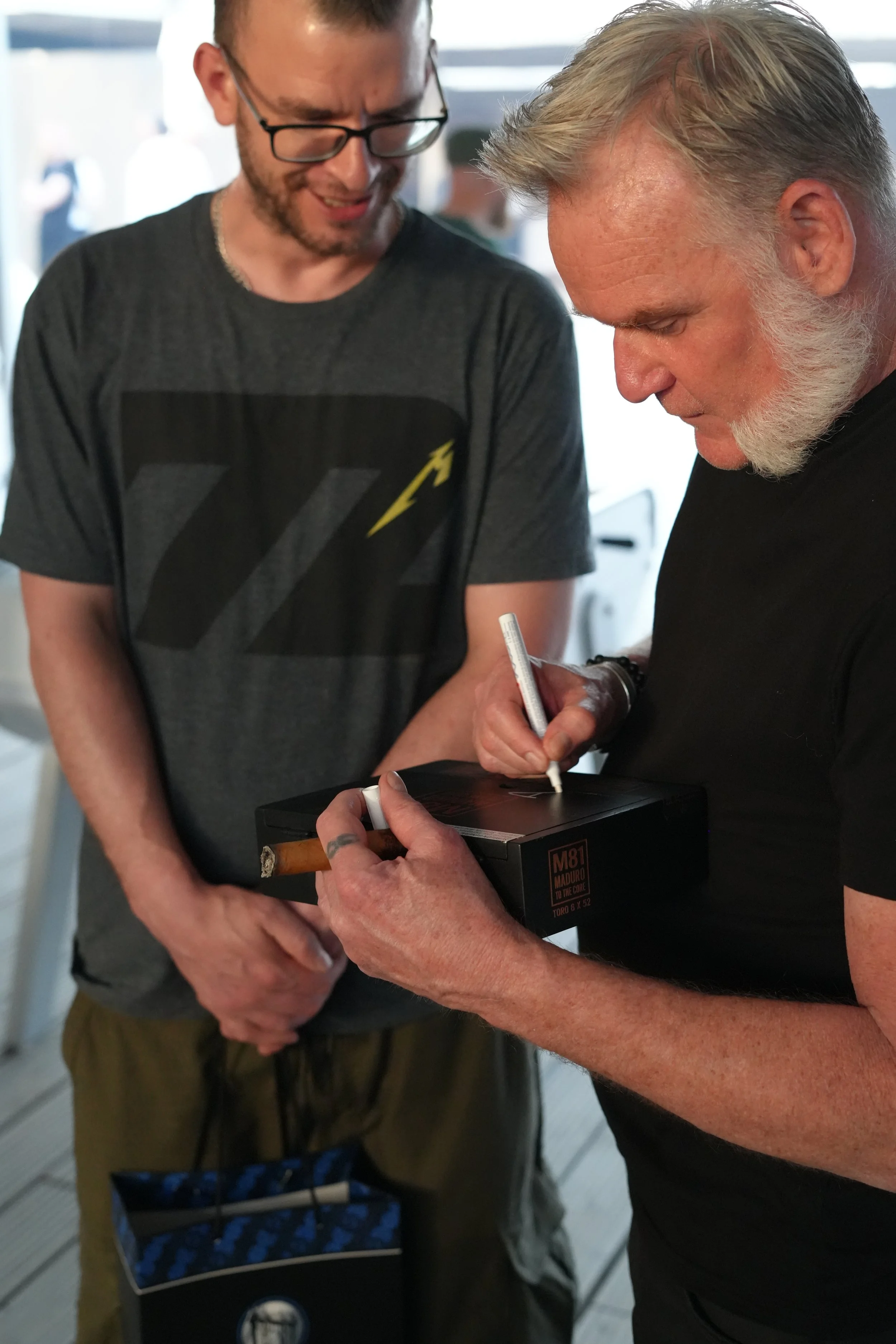 Two men are in a conversation. One is signing an item, and the other is watching and smiling. The signer has a beard and gray hair. The other man wears glasses. They are indoors, with a blurred background and a shopping basket visible.