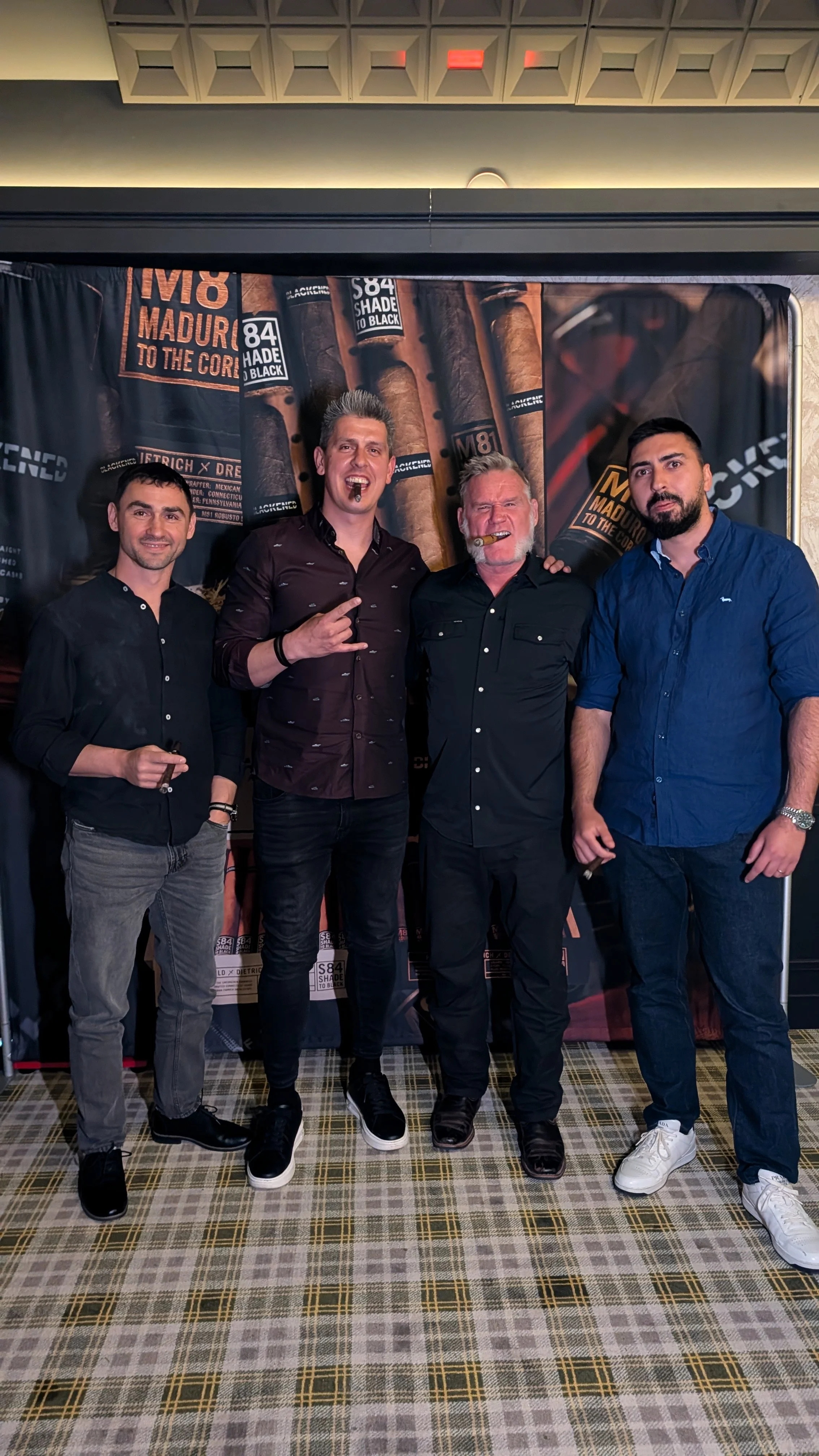 Four men standing together inside a room with a large cigar-themed backdrop. The men are dressed casually, with three of them holding cigars and smiling or making hand gestures.