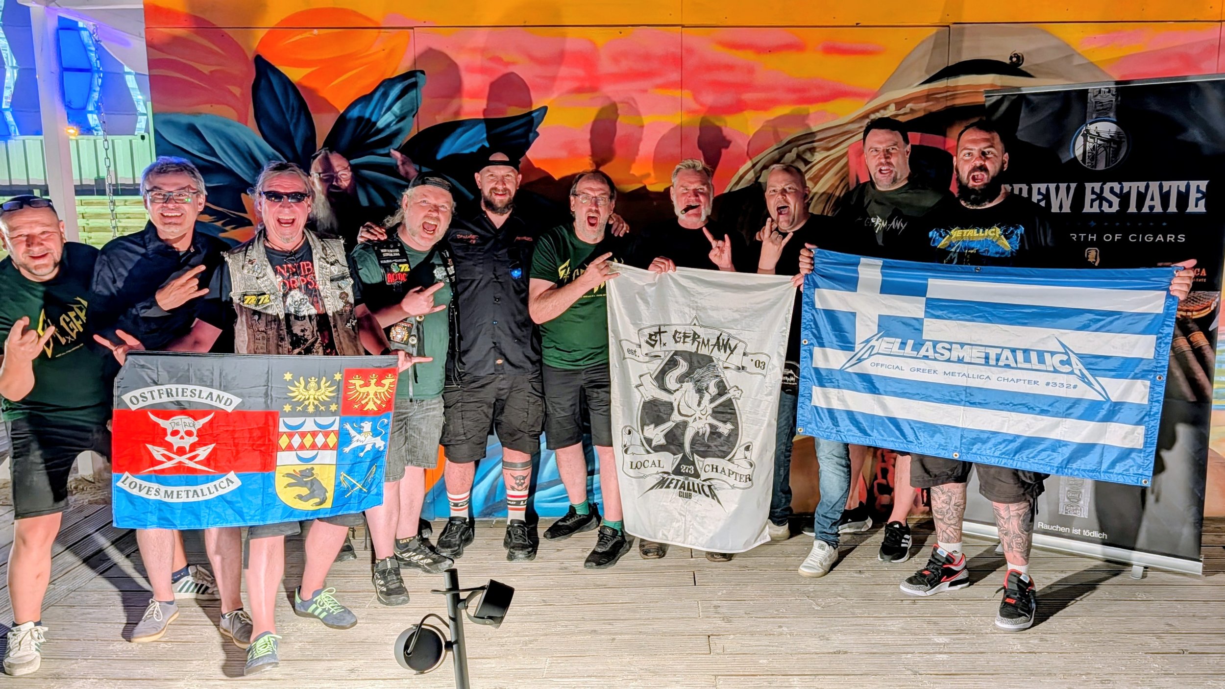 A group of men posing with Metallica chapter flags in front of a colorful mural with Rob Dietrich in Frankfurt, Germany.