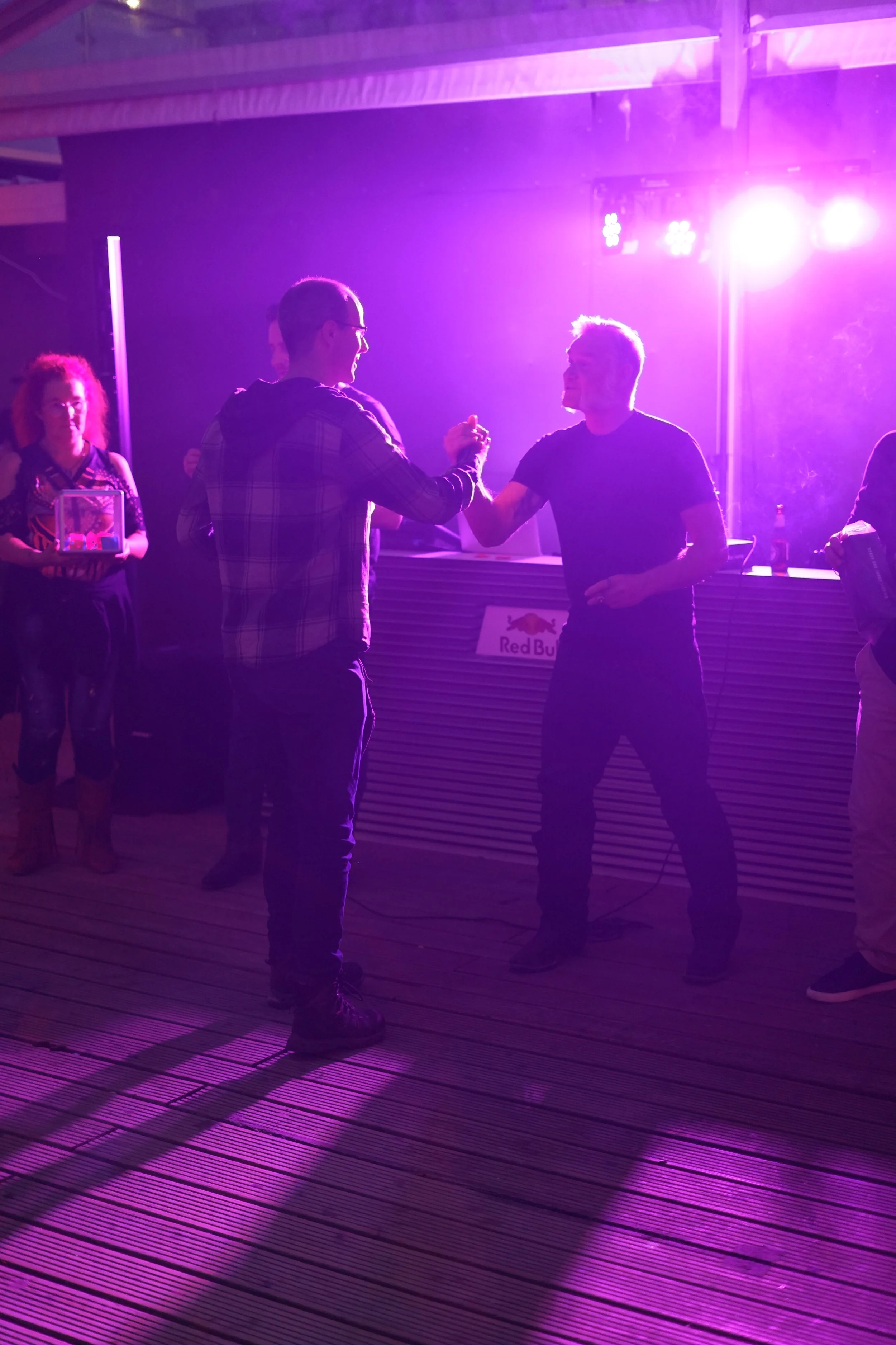 Two men in dark clothing engaging in a handshake, illuminated by purple stage lighting, with a woman in the background holding a small box, at an event with a Red Bull sign and a DJ setup.