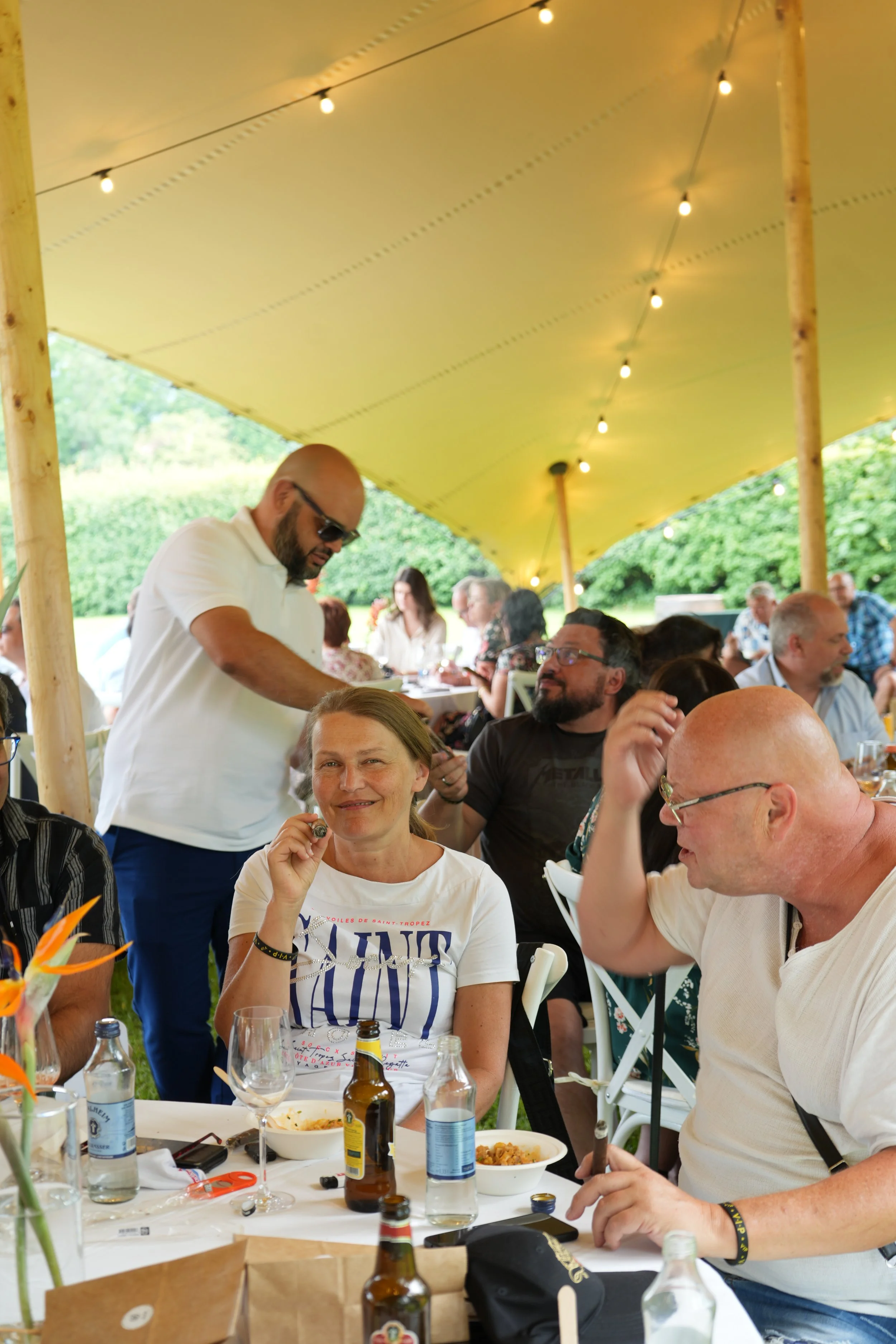 People gathered at a social outdoor event under a yellow canopy with string lights, enjoying food and drinks at a long table.