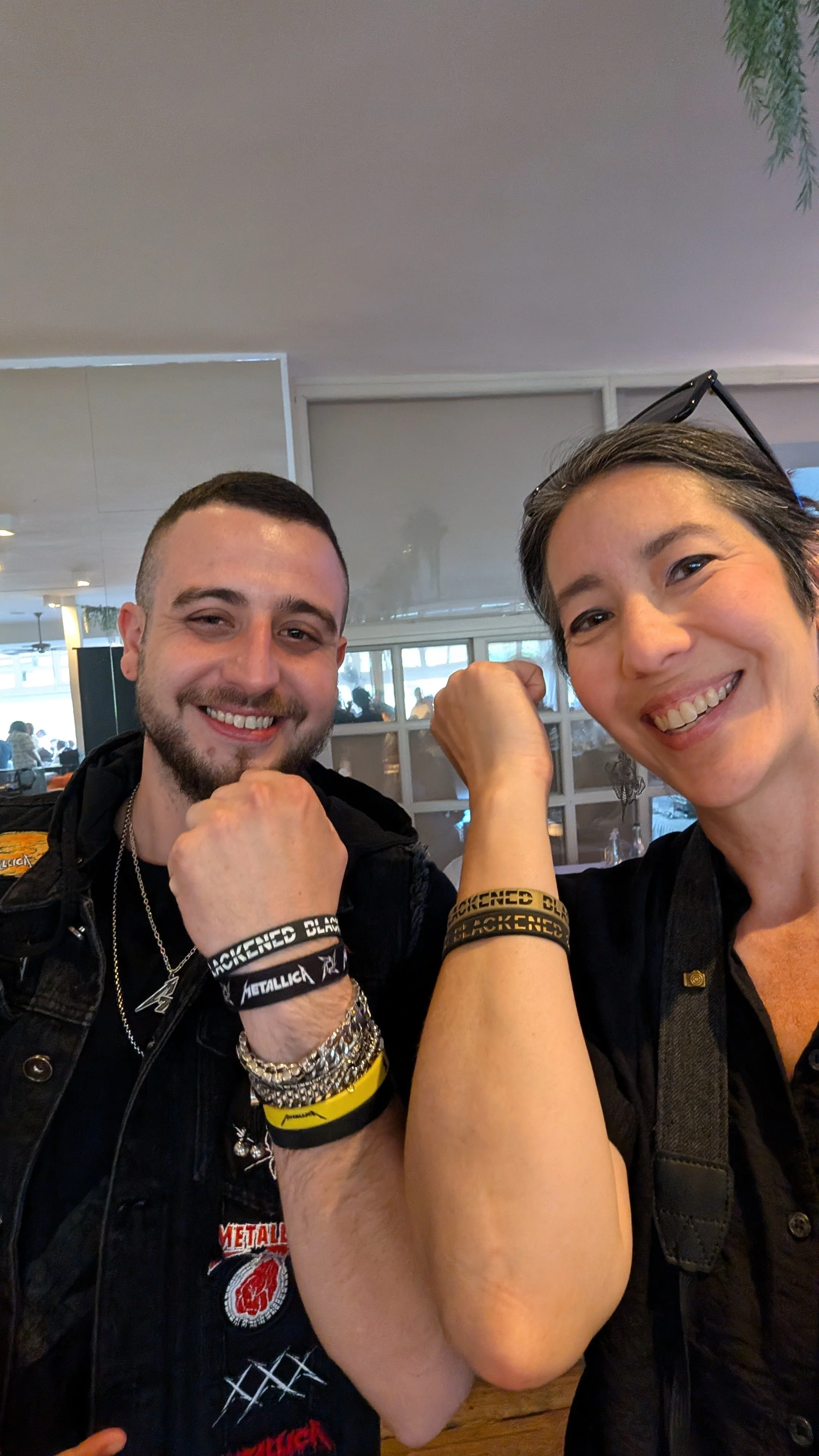 Two smiling women, flexing their arms and showing off their bracelets, in a restaurant or cafe setting.
