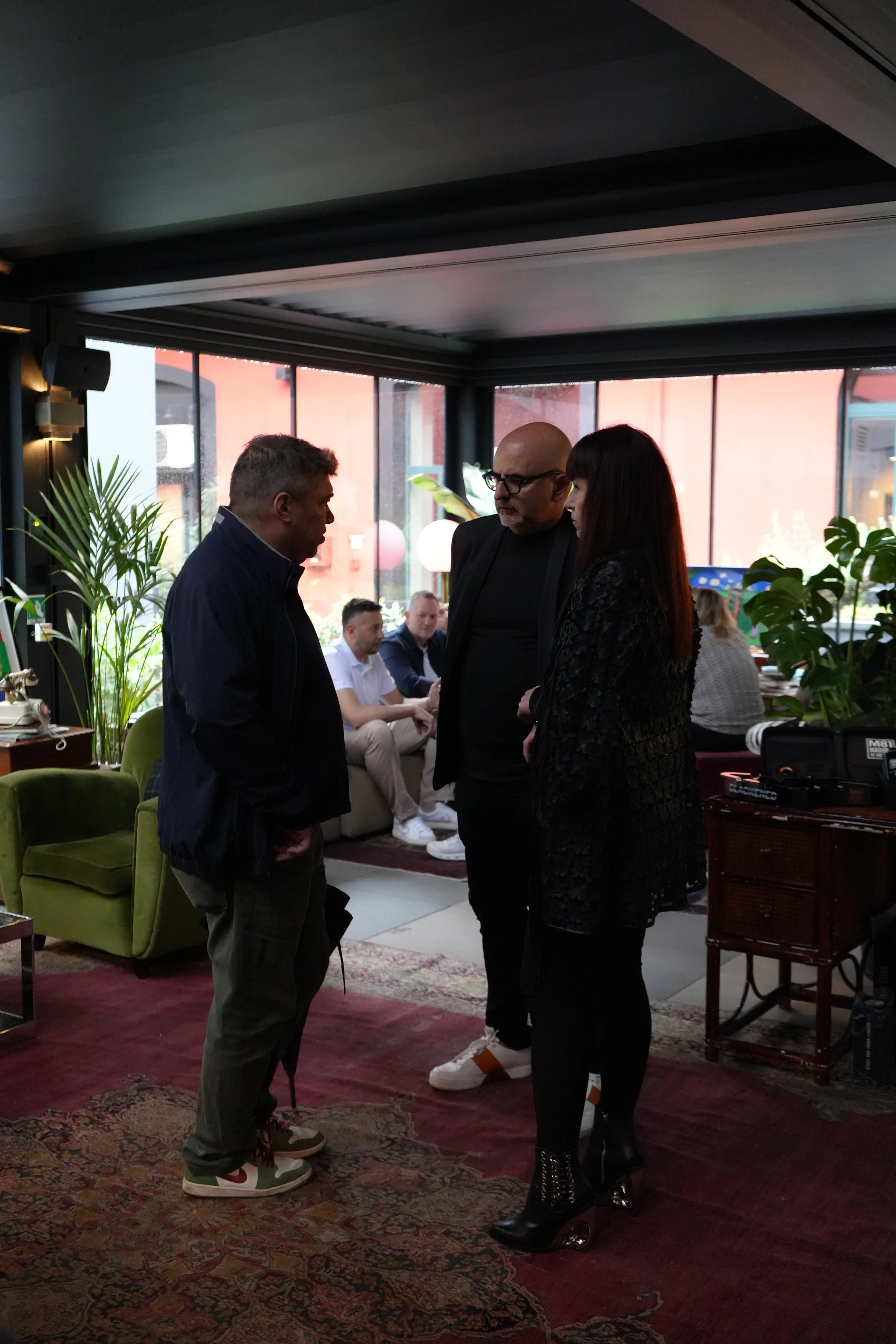 Three people, two men and one woman, are having a conversation in a cozy, indoor cafe or restaurant setting with large windows, plants, and comfortable furniture. The woman is wearing black clothing and boots, while the men are dressed casually.
