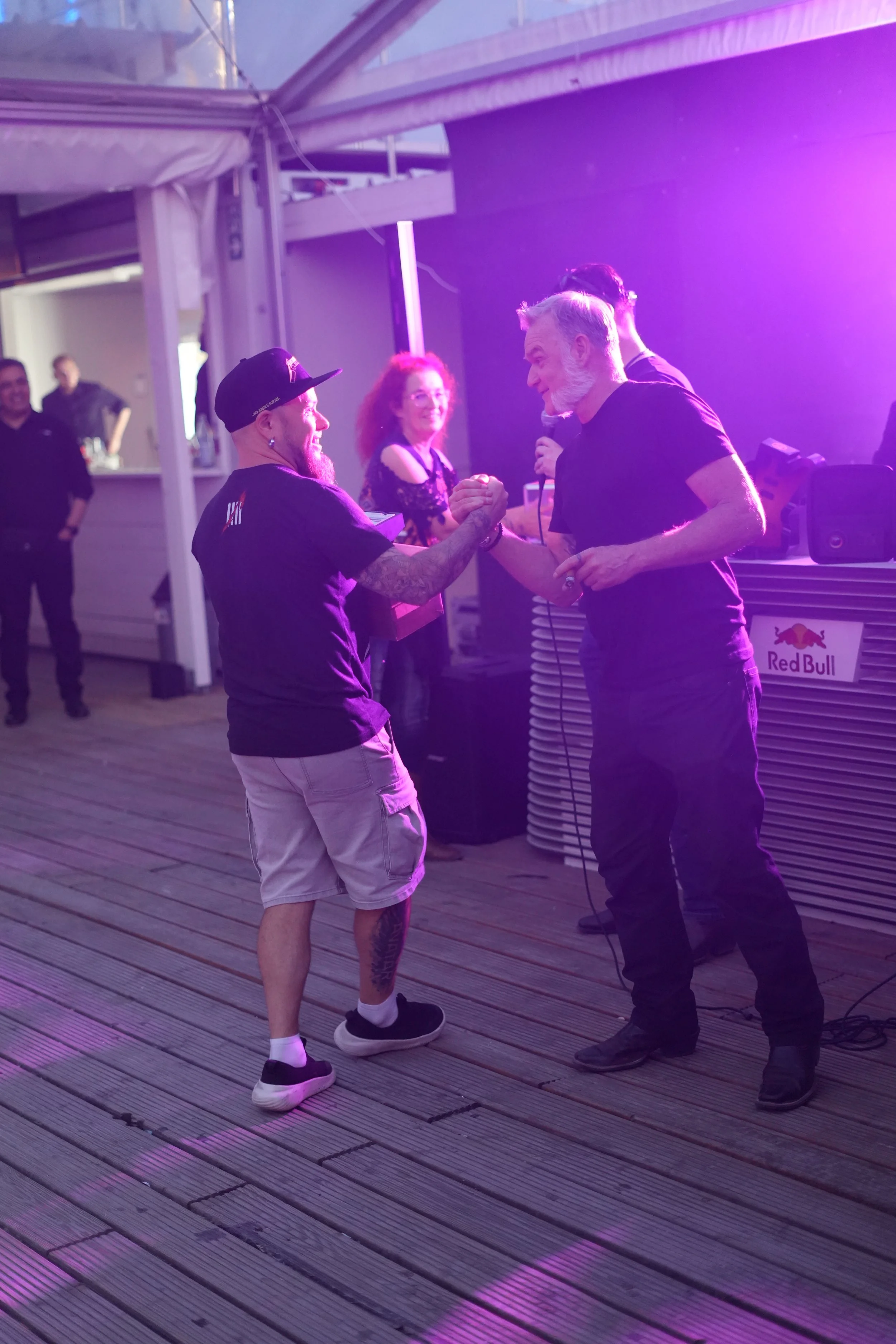 Two men smiling and shaking hands at a party or event, with a woman in the background holding a microphone and a DJ booth with a Red Bull logo, pink and purple lighting.
