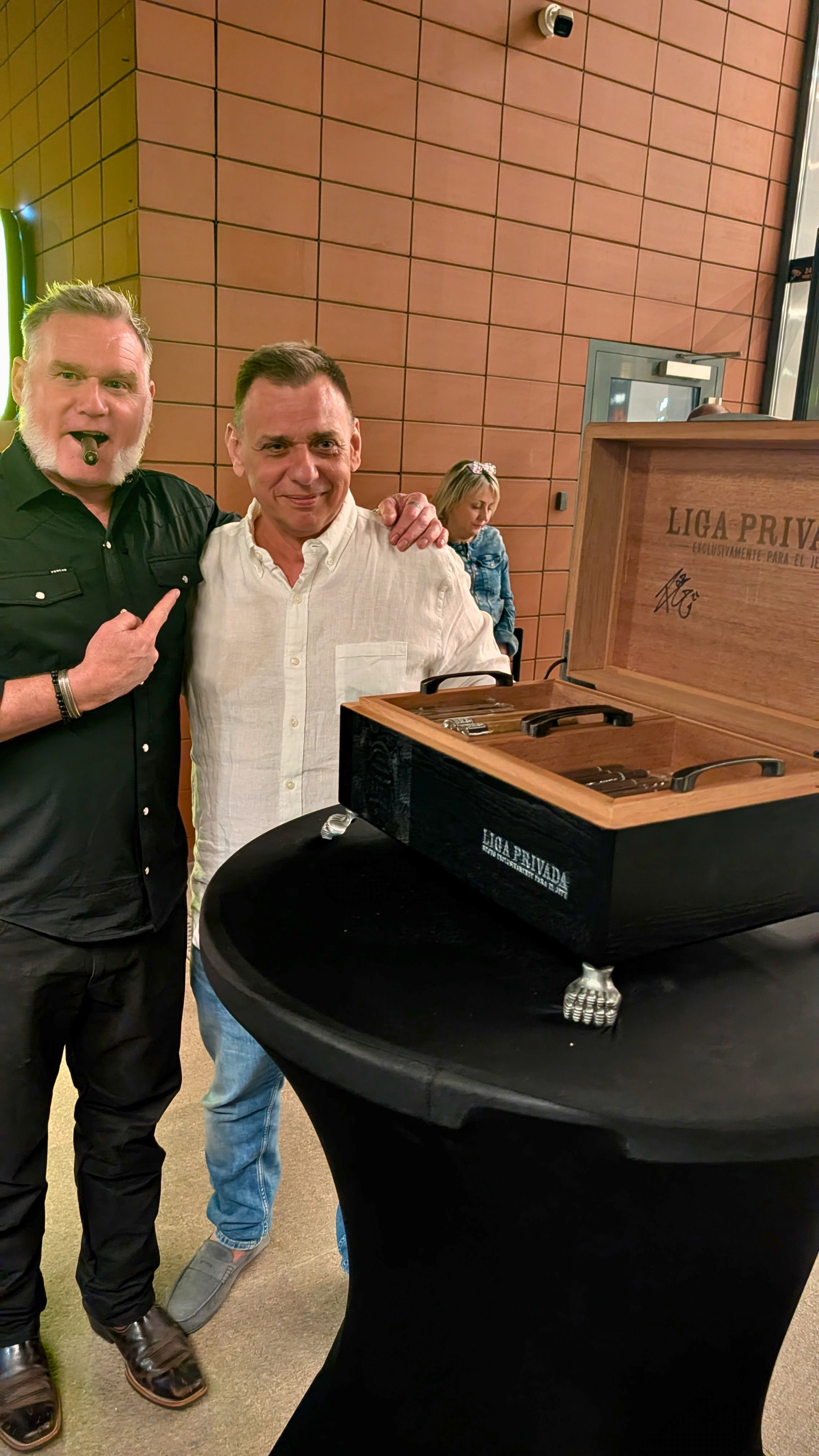 Two men are standing next to a table with a cigar humidor, and an overworked man is making a gesture with his finger. The background features a brick wall and a woman sitting in a corner.