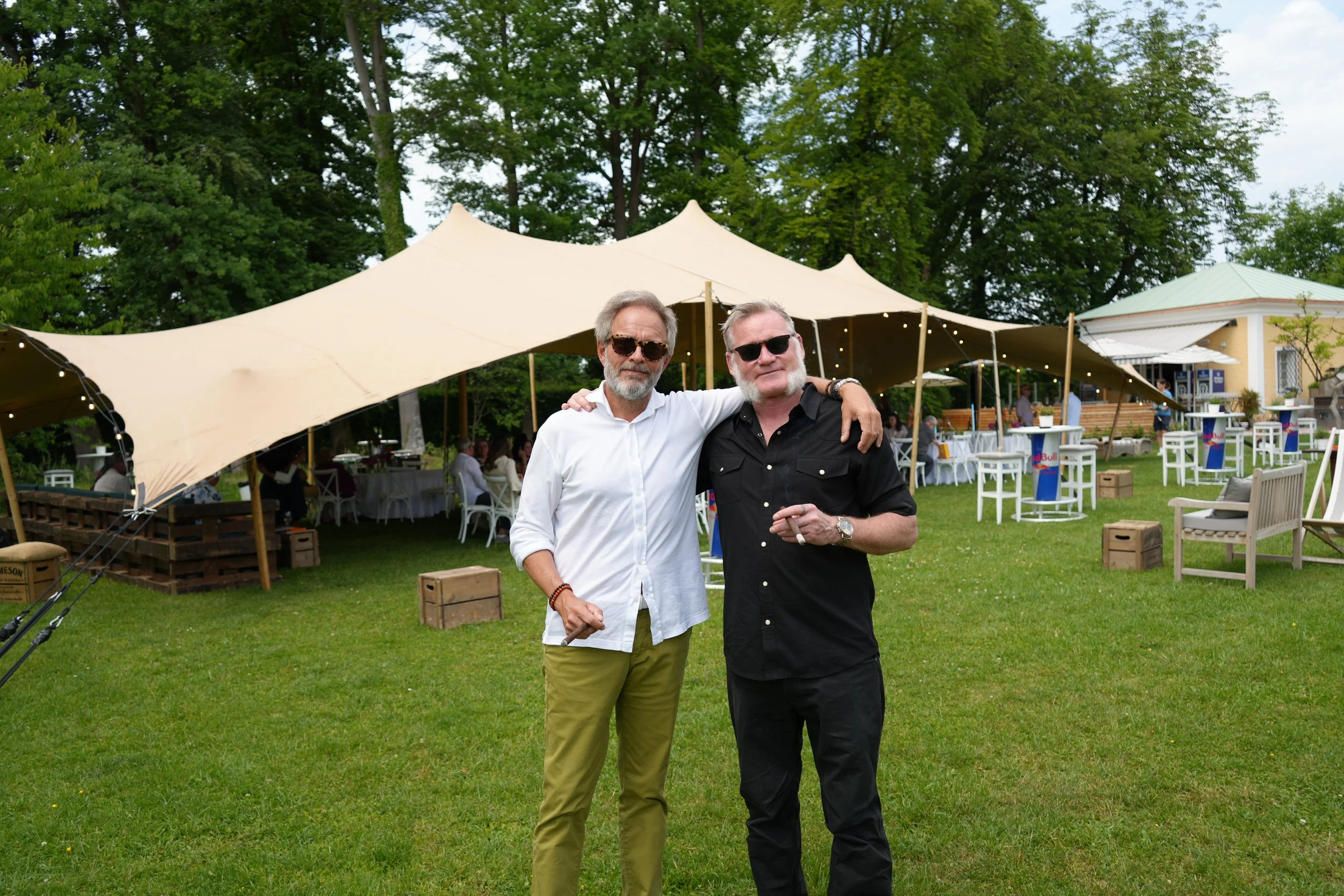 Two men standing together at an outdoor event, one in a white shirt and khaki pants, the other in a black shirt and black pants. They are wearing sunglasses and are smiling, with their arms around each other. Behind them is a large beige tent, trees,