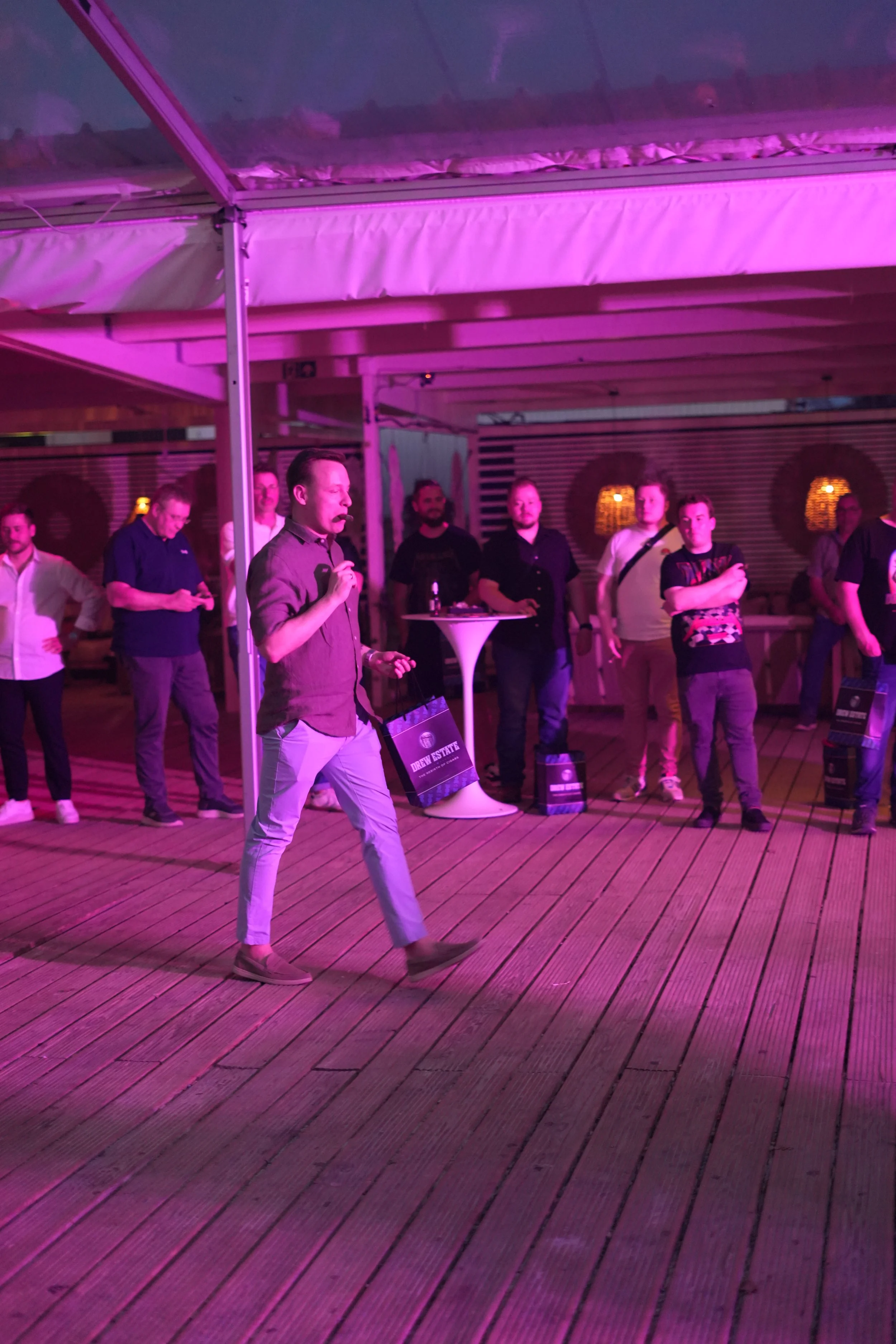 A man speaking or singing into a microphone on a wooden deck under purple lighting, with several people standing in the background.