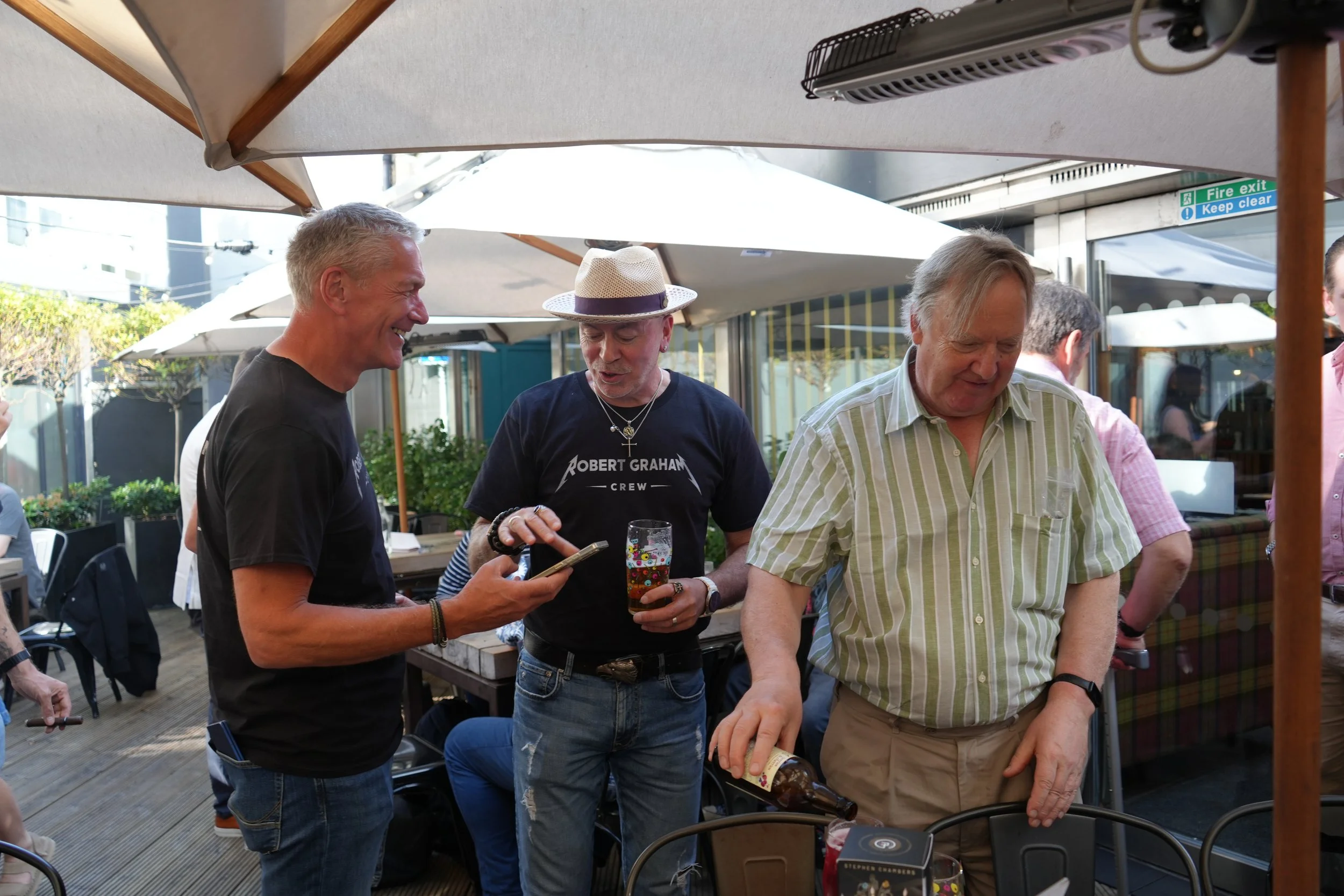 A group of men socializing outdoors under umbrellas, some holding drinks, engaging in conversation during daytime.