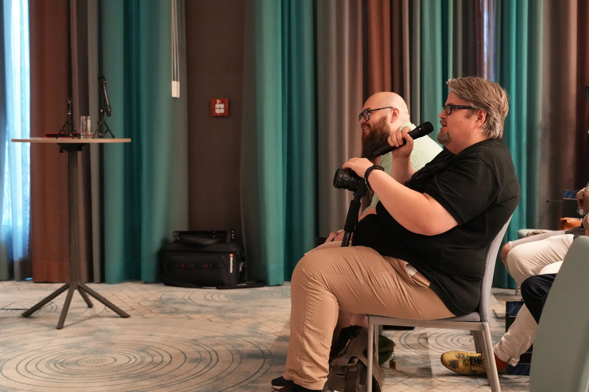 Two men sitting and smiling at a conference or seminar, one holding a microphone. There are curtains and a small table with equipment in the background.