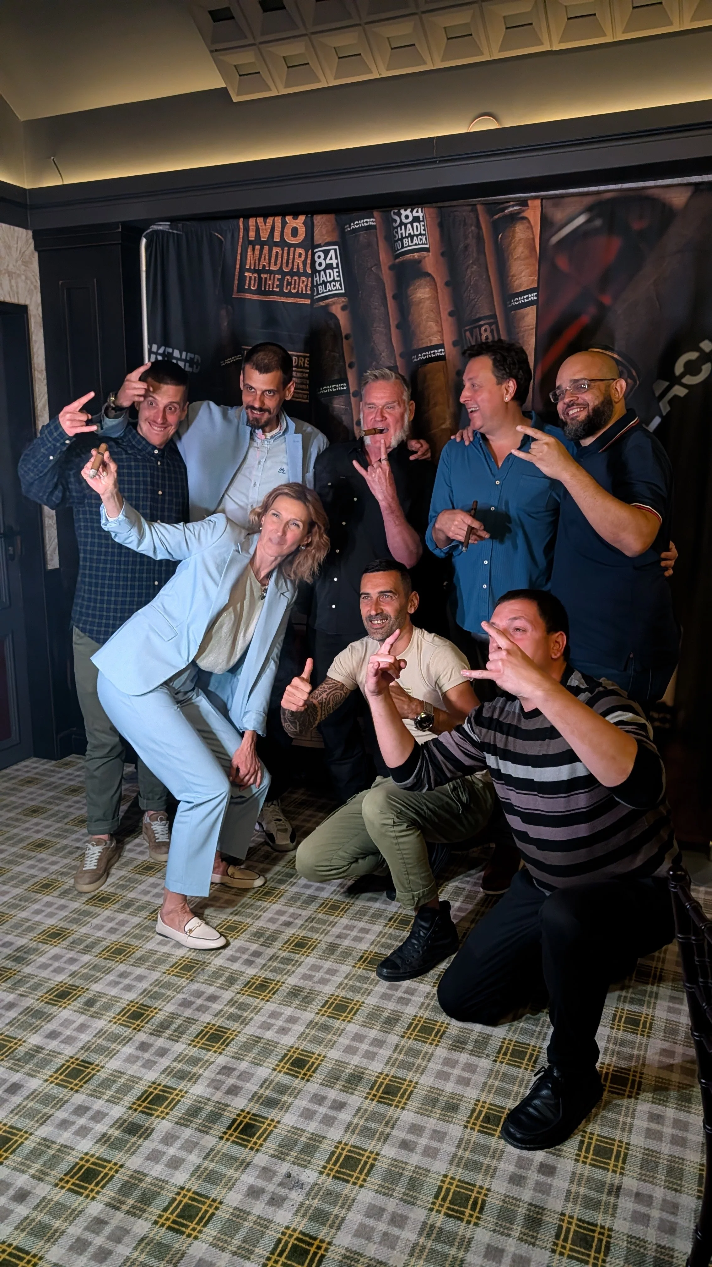 Group of nine people posing and making hand gestures in front of a backdrop featuring cigars and cigar brand names.
