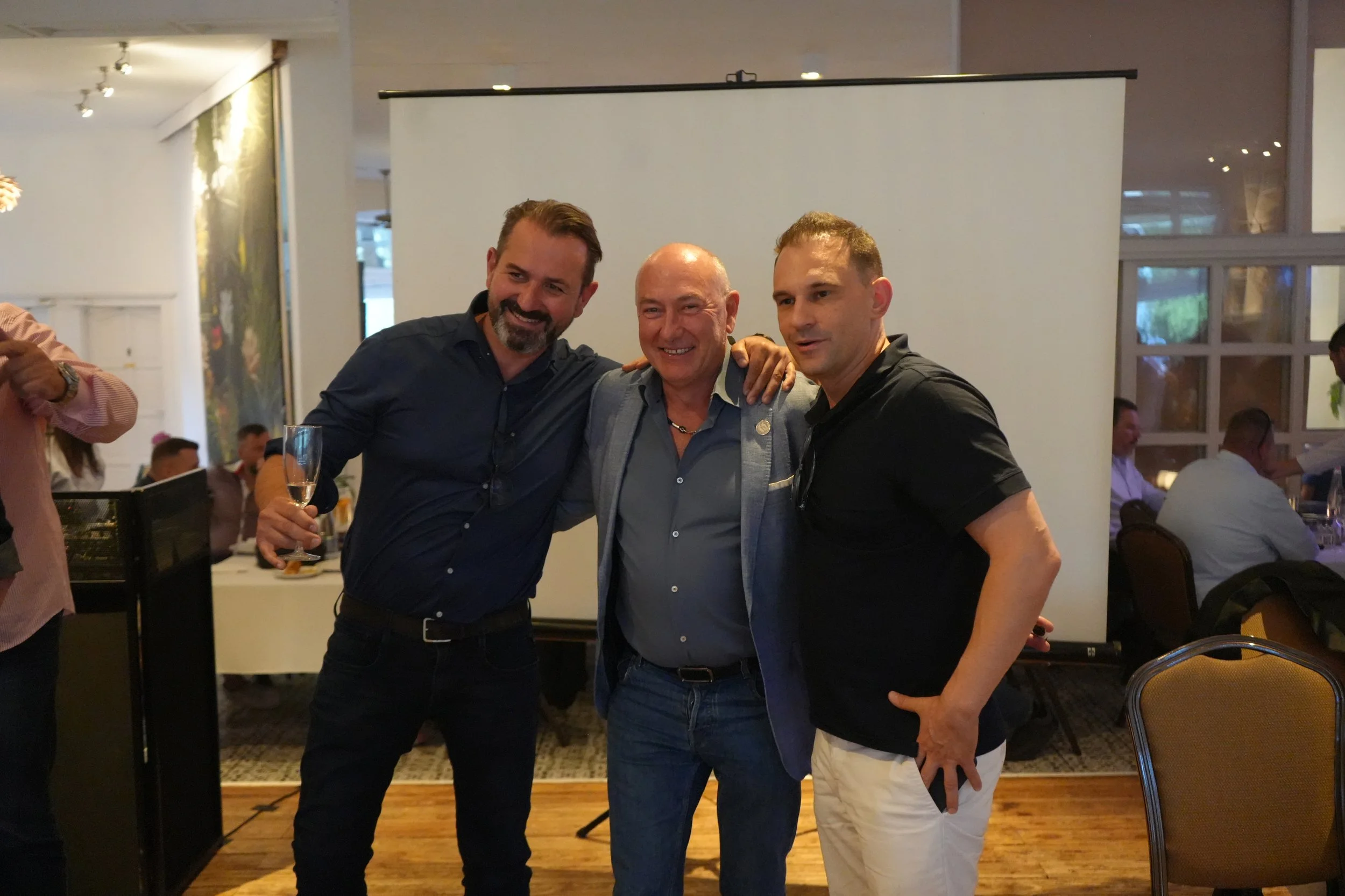 Three men standing together indoors at a social event, smiling at the camera. The man on the left holds a drink, the middle man is bald, and the man on the right is wearing a black shirt.