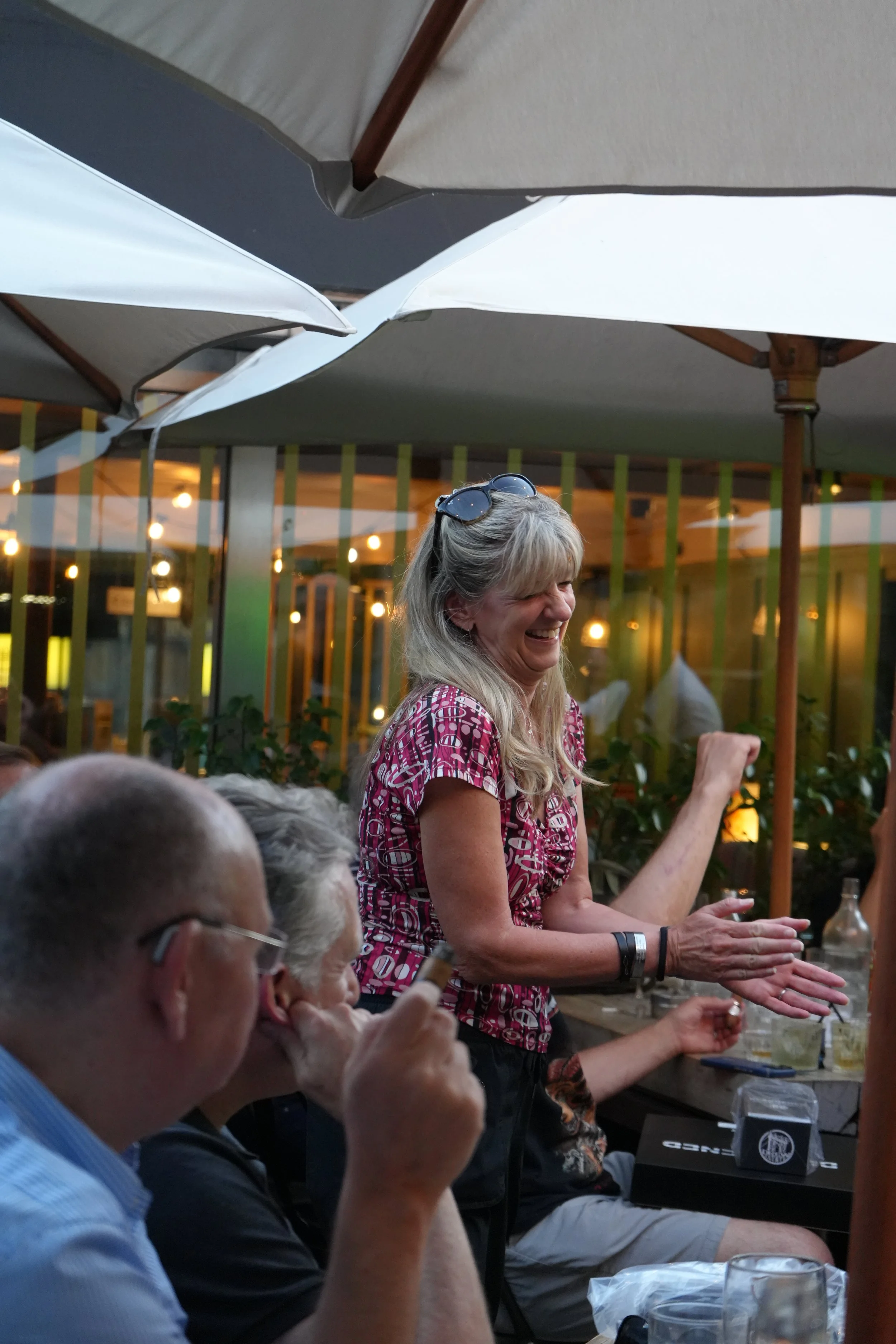 A woman with blonde hair and sunglasses on her head is smiling and clapping while standing among seated people at an outdoor gathering under umbrellas.