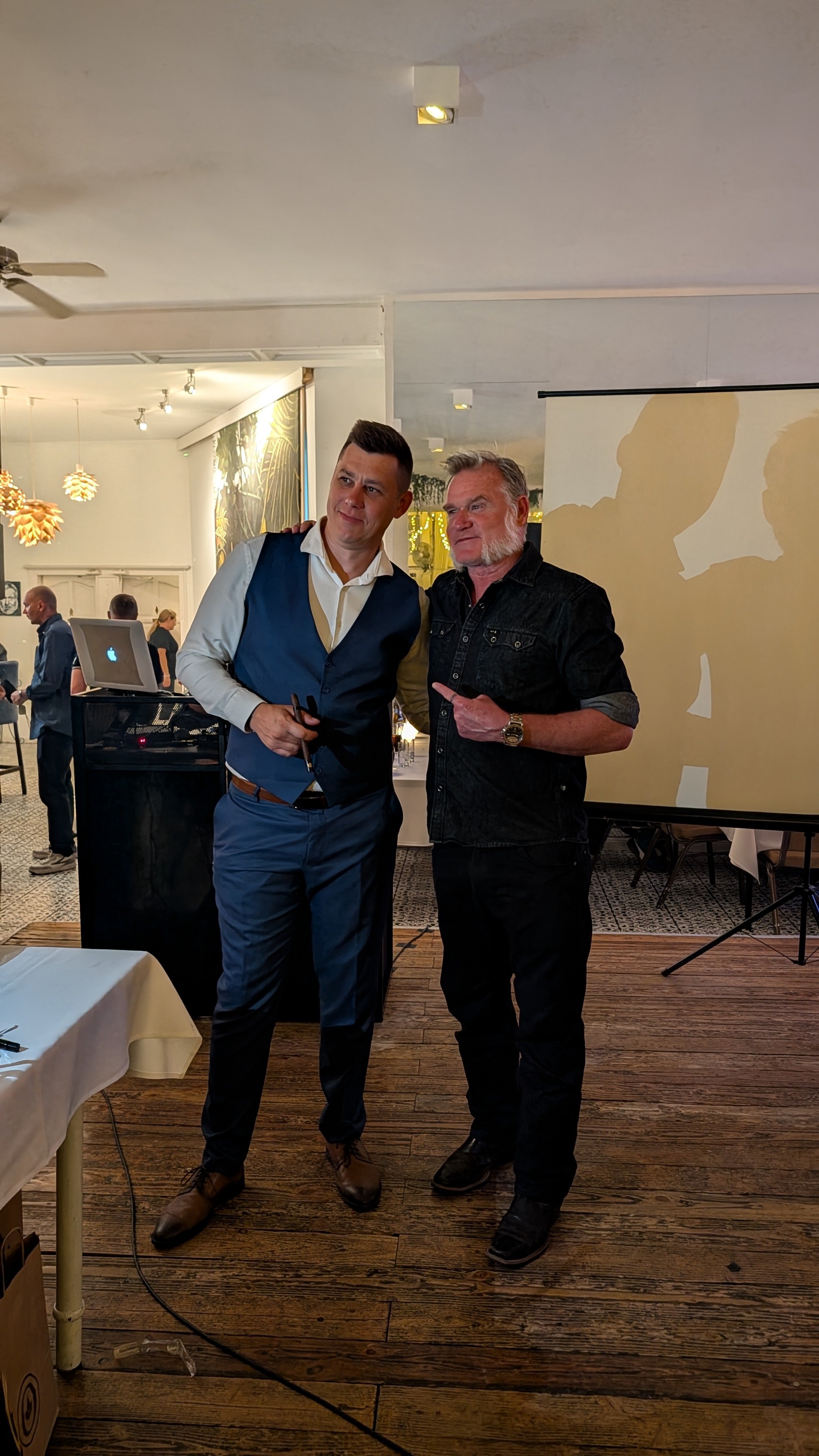 Two men stand together in a restaurant or event space. One man is dressed in a blue vest and white shirt, holding glasses, while the other man is in black attire and pointing at the first. They pose for the photo in front of a large screen and variou