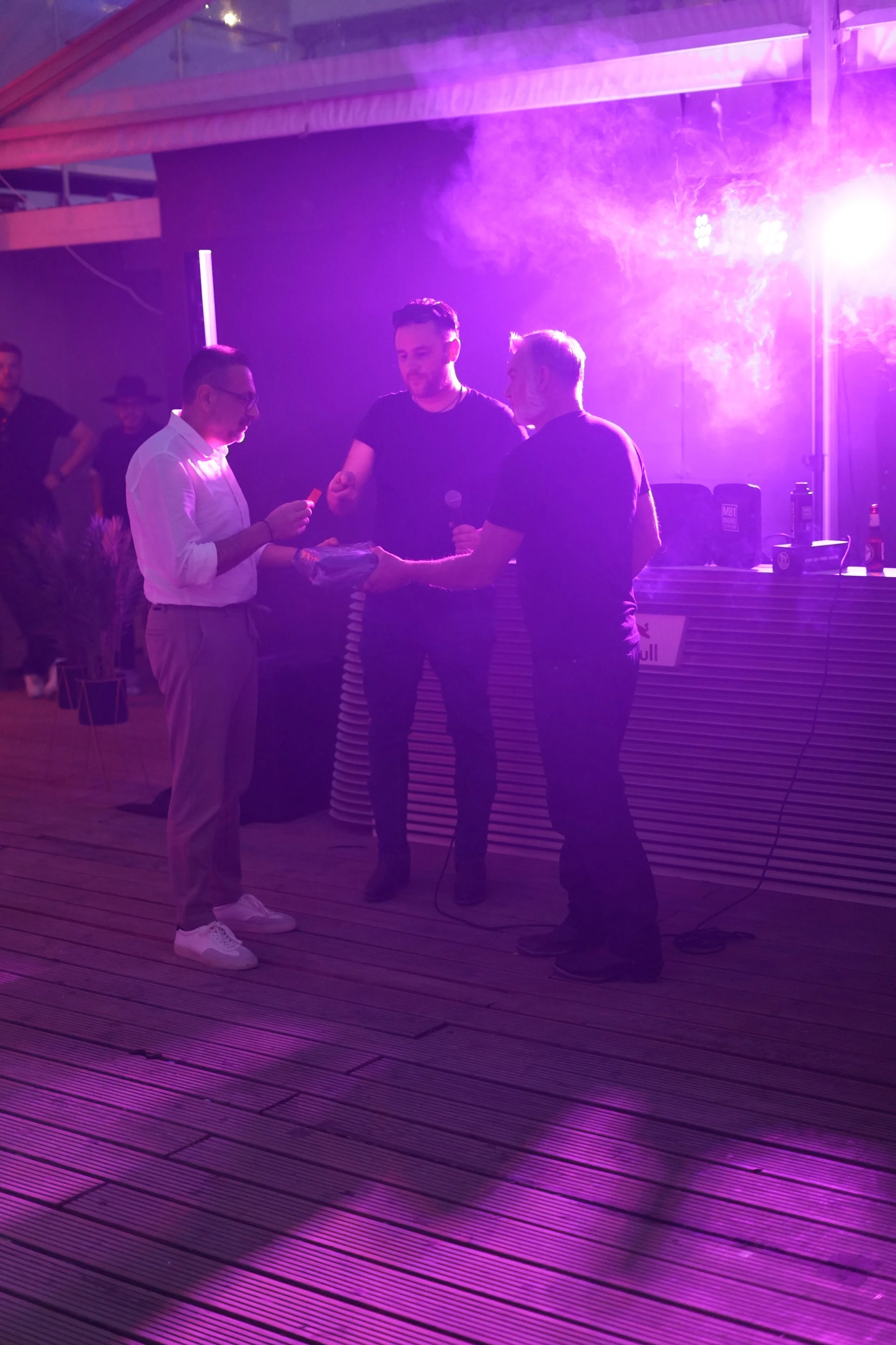 Three men are standing and talking in a venue with purple and pink lighting. One man is holding some papers, another is holding a microphone, and the third is handing over a gift or item. There are some bottles and equipment on a counter behind them.