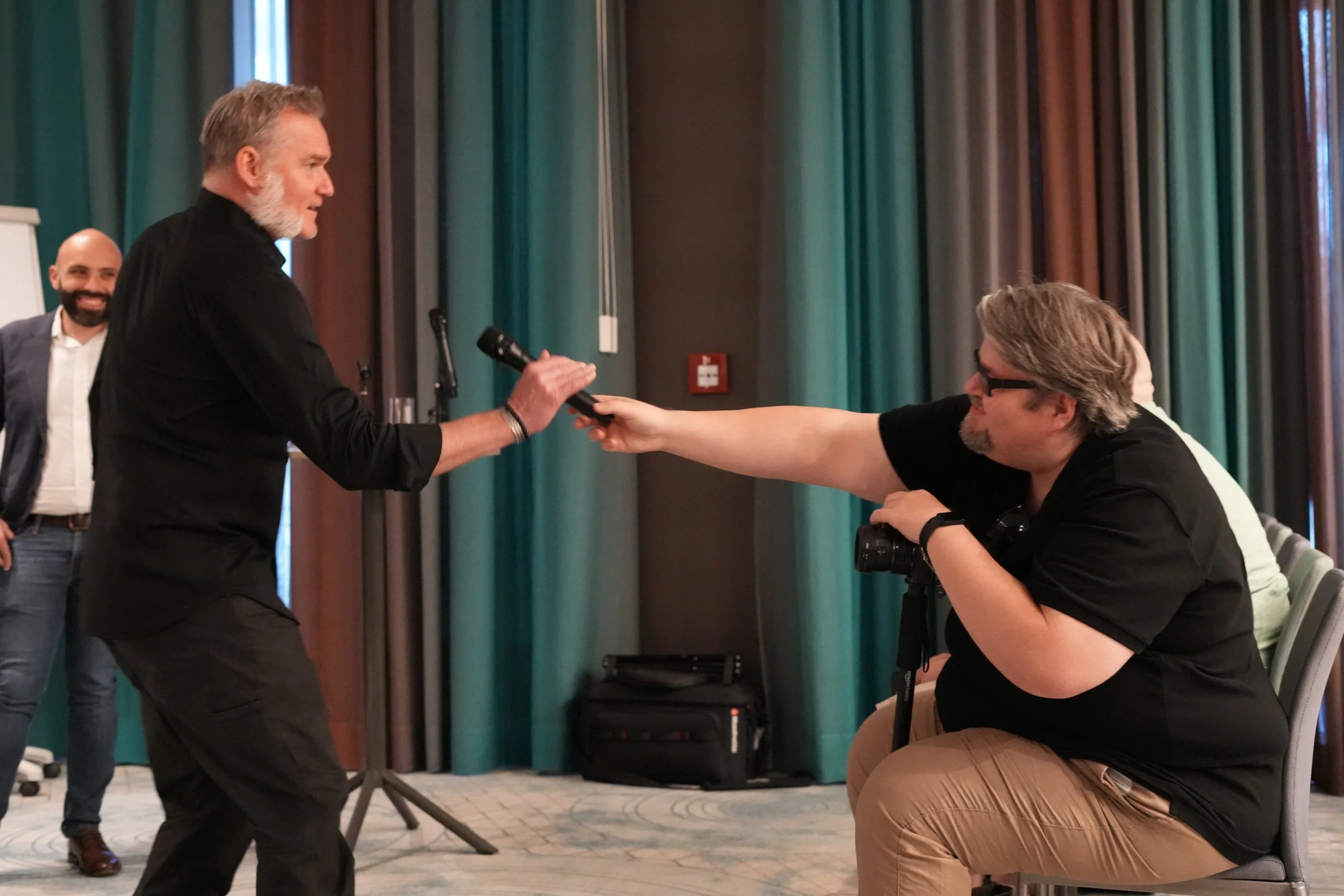 A man with gray hair and beard holding a microphone in a conference room, interviewing a seated man with glasses, gray hair, and a camera around his neck, while another man in the background smiles.