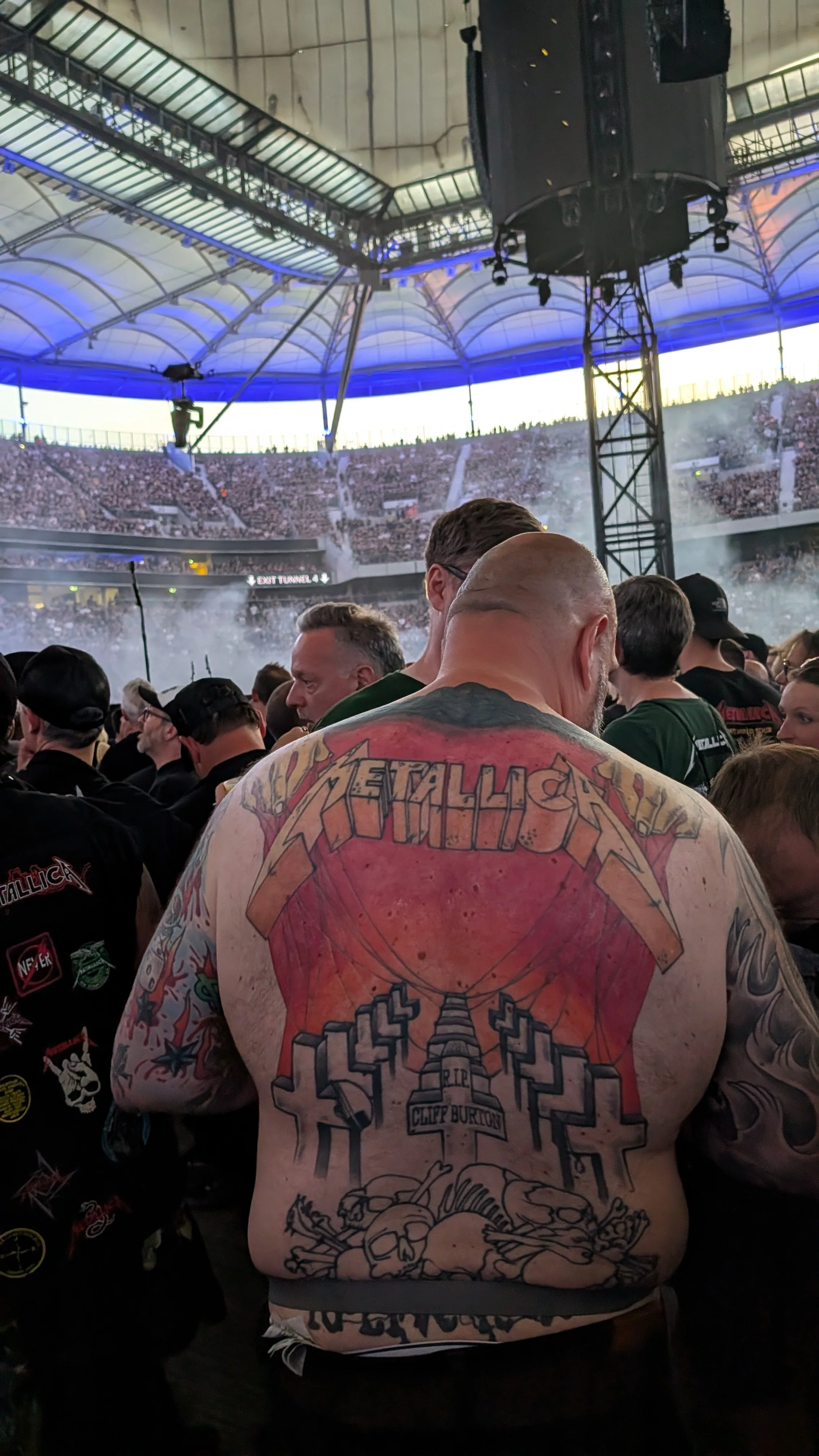 Crowd inside a stadium for a Metallica concert or event with a large speaker hanging from the ceiling and a man with extensive colorful tattoos on his back in the foreground.