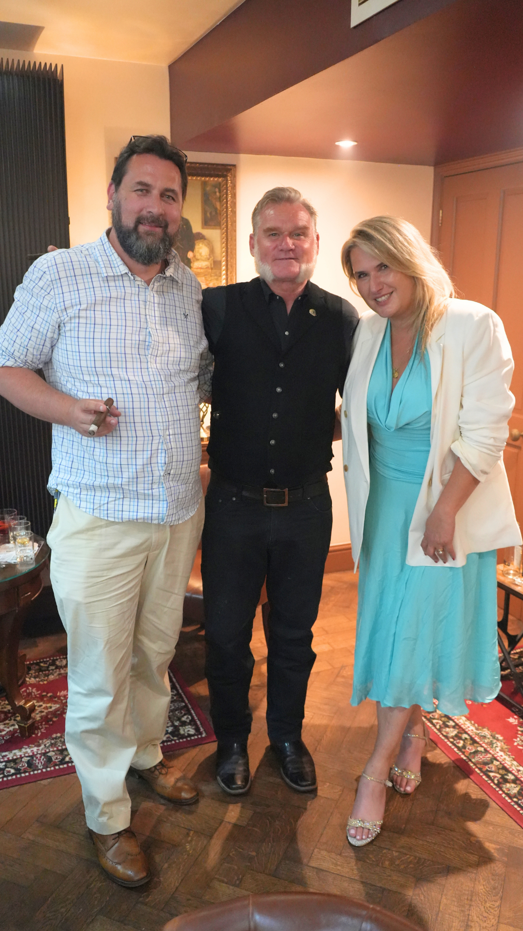 Three people standing together in a room, smiling. The man on the left is wearing a checkered shirt, light-colored pants, and brown shoes. The man in the middle is dressed in black with a black vest and black pants. The woman on the right is wearing 