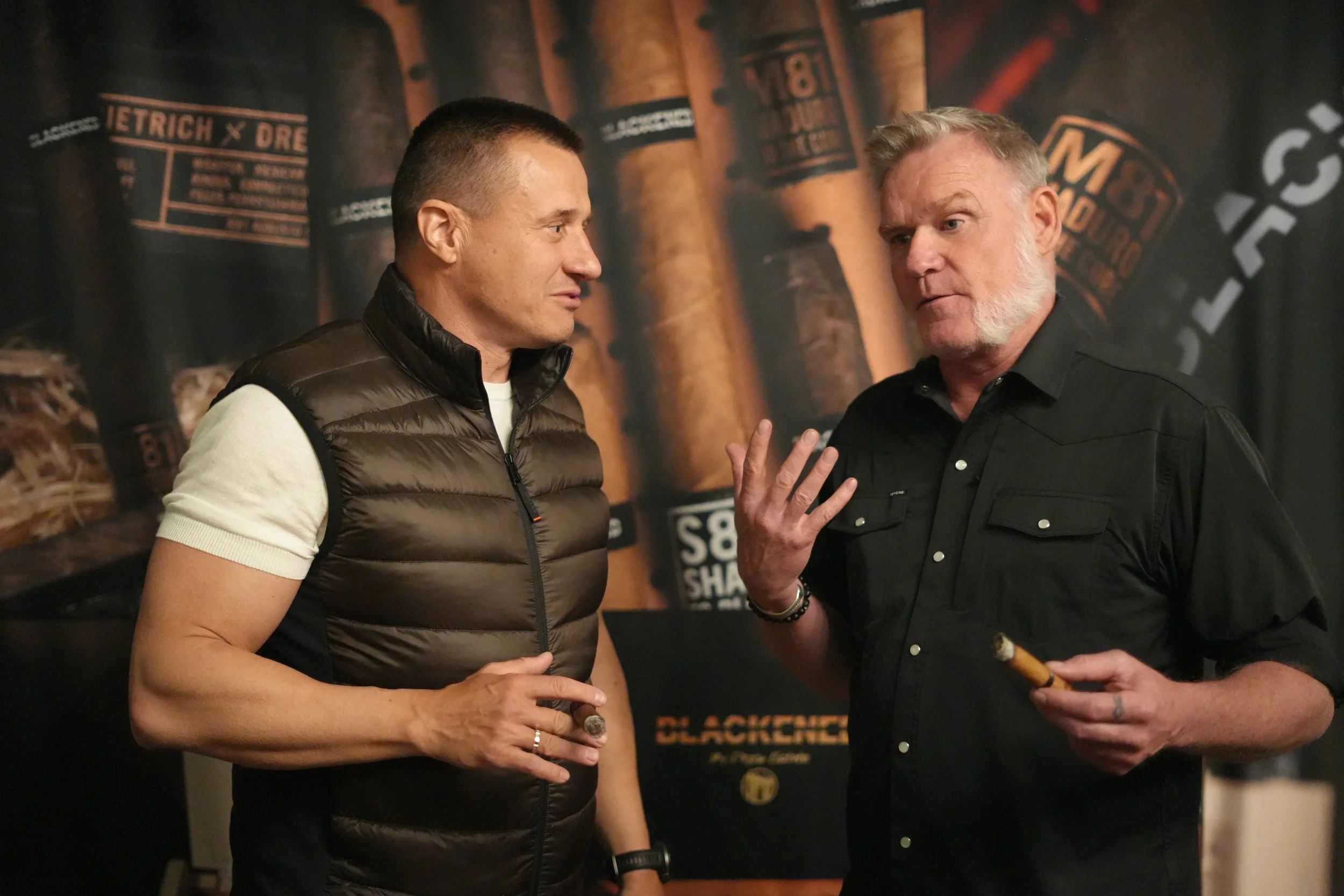 Two men engaged in conversation at an event, with a dark background featuring shelving and promotional posters related to cigars.