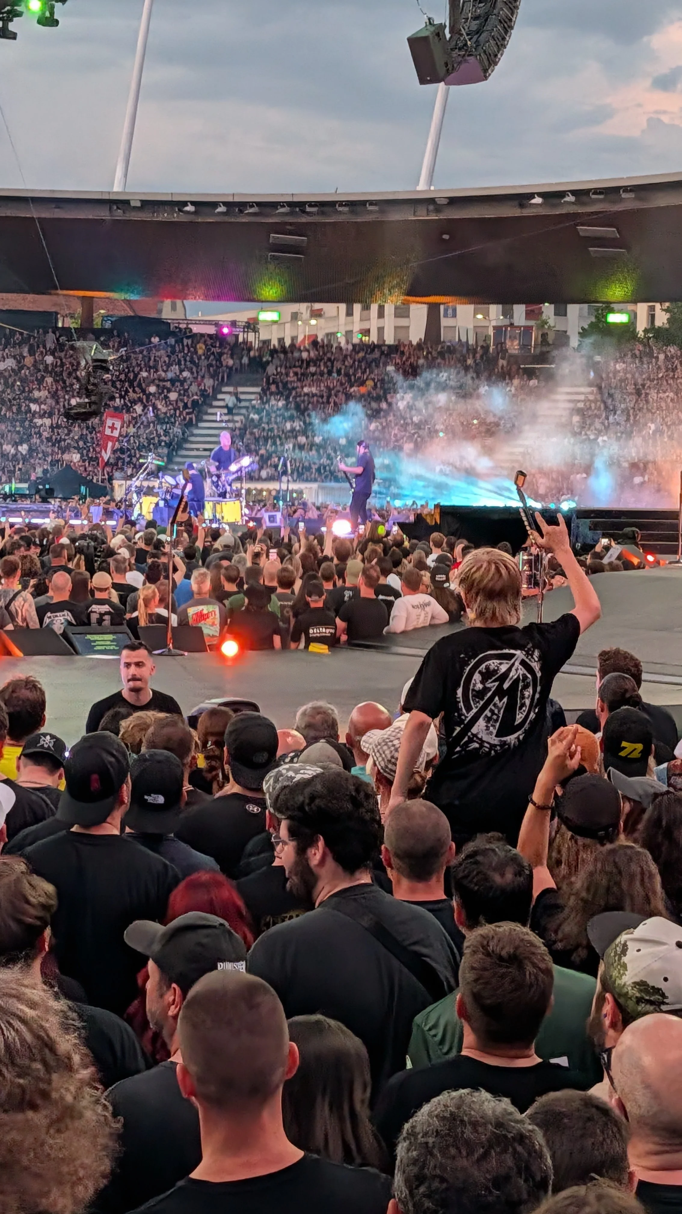 Crowd watching a Metallica concert, with stage and performers in the background, and colorful smoke and lighting effects.