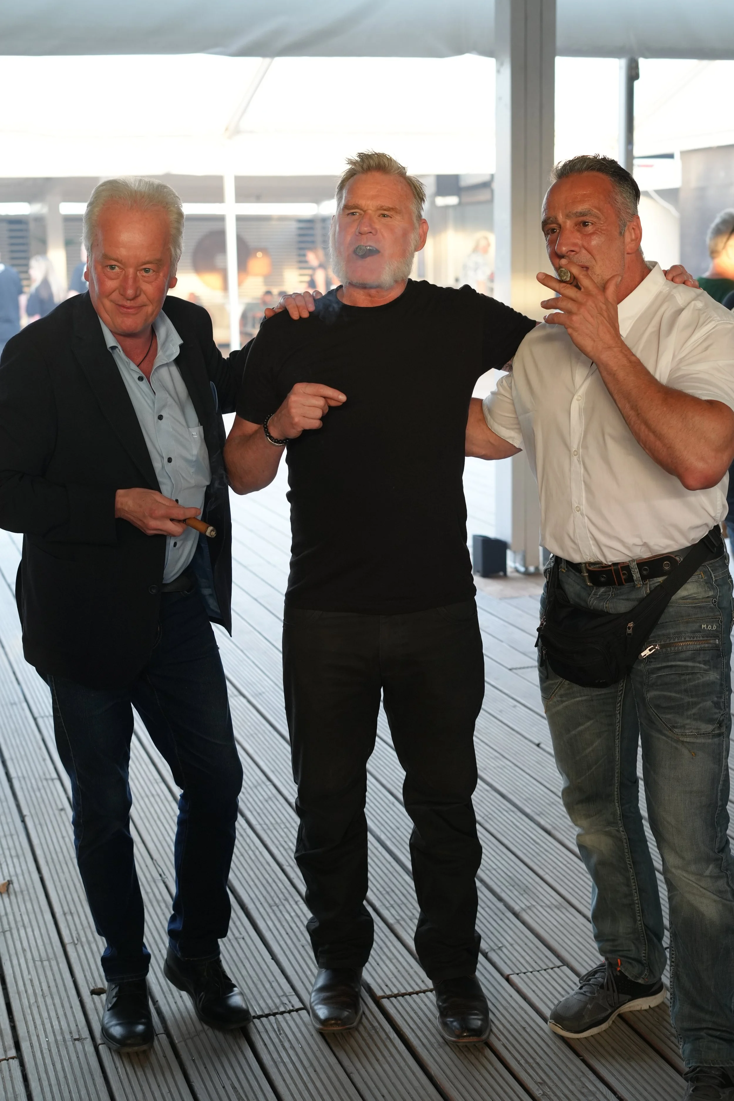 Three middle-aged men standing together on a wooden deck, arms around each other, smoking cigars, with a glass enclosure and outdoor background.
