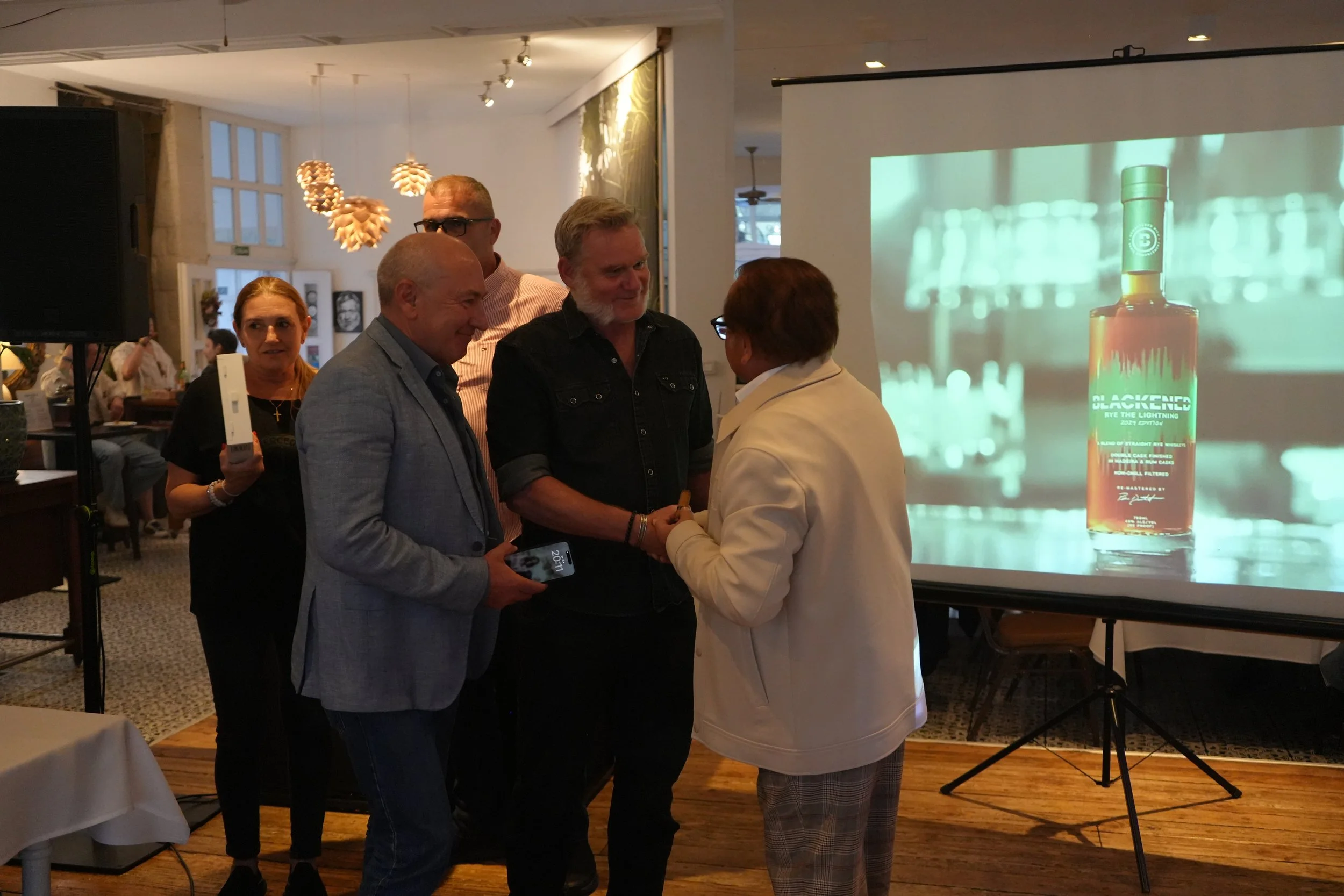 A group of five people engaged in conversation at a social event, with a man and woman shaking hands and others smiling nearby, in a room decorated with distinctive hanging light fixtures and a large screen displaying a product advertisement.