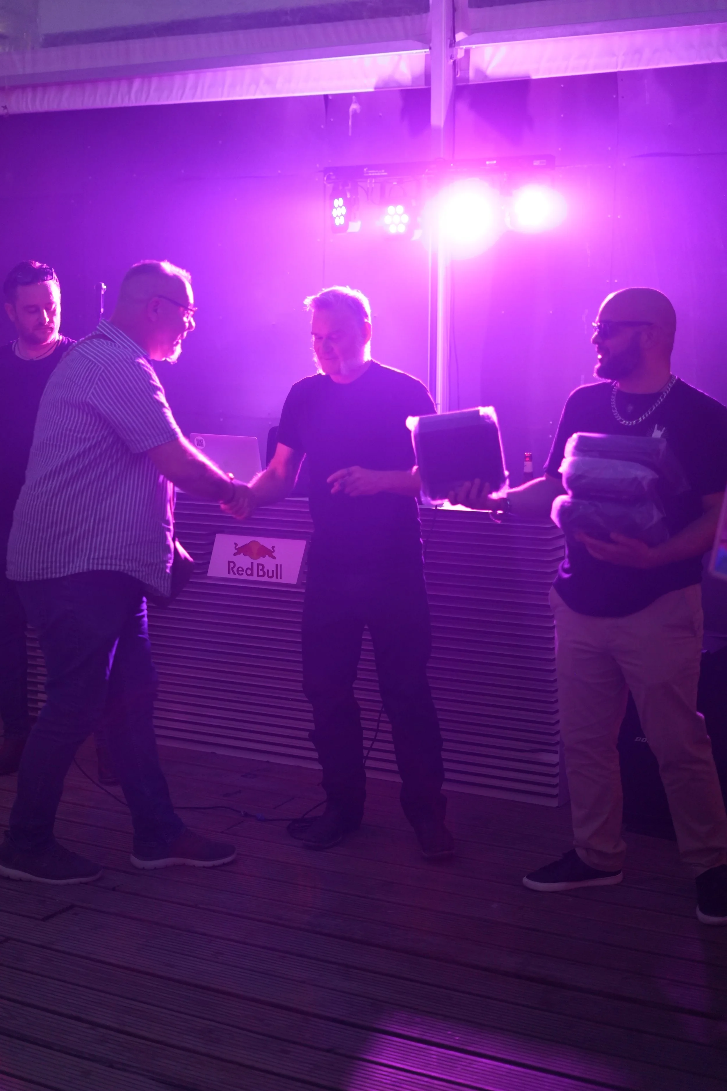 Four men at a nightclub or event under purple lights, exchanging a handshake and holding a folder, with a Red Bull sign in the background.