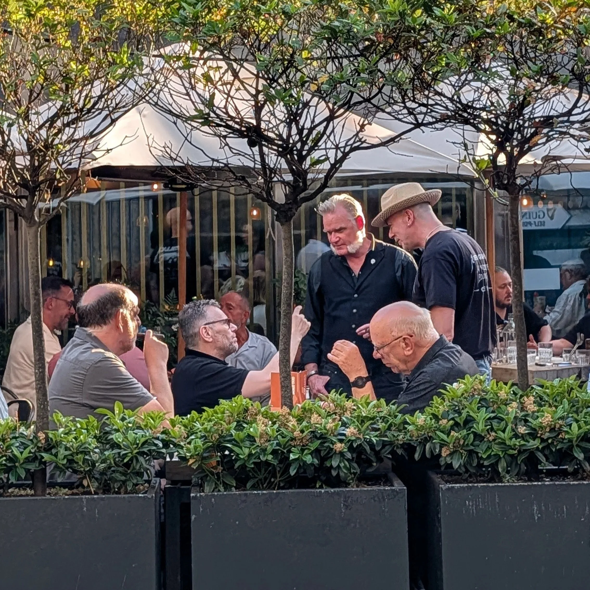 People sitting outdoors at a restaurant or bar, with a tree in the foreground and string lights hanging above, engaging in conversation.