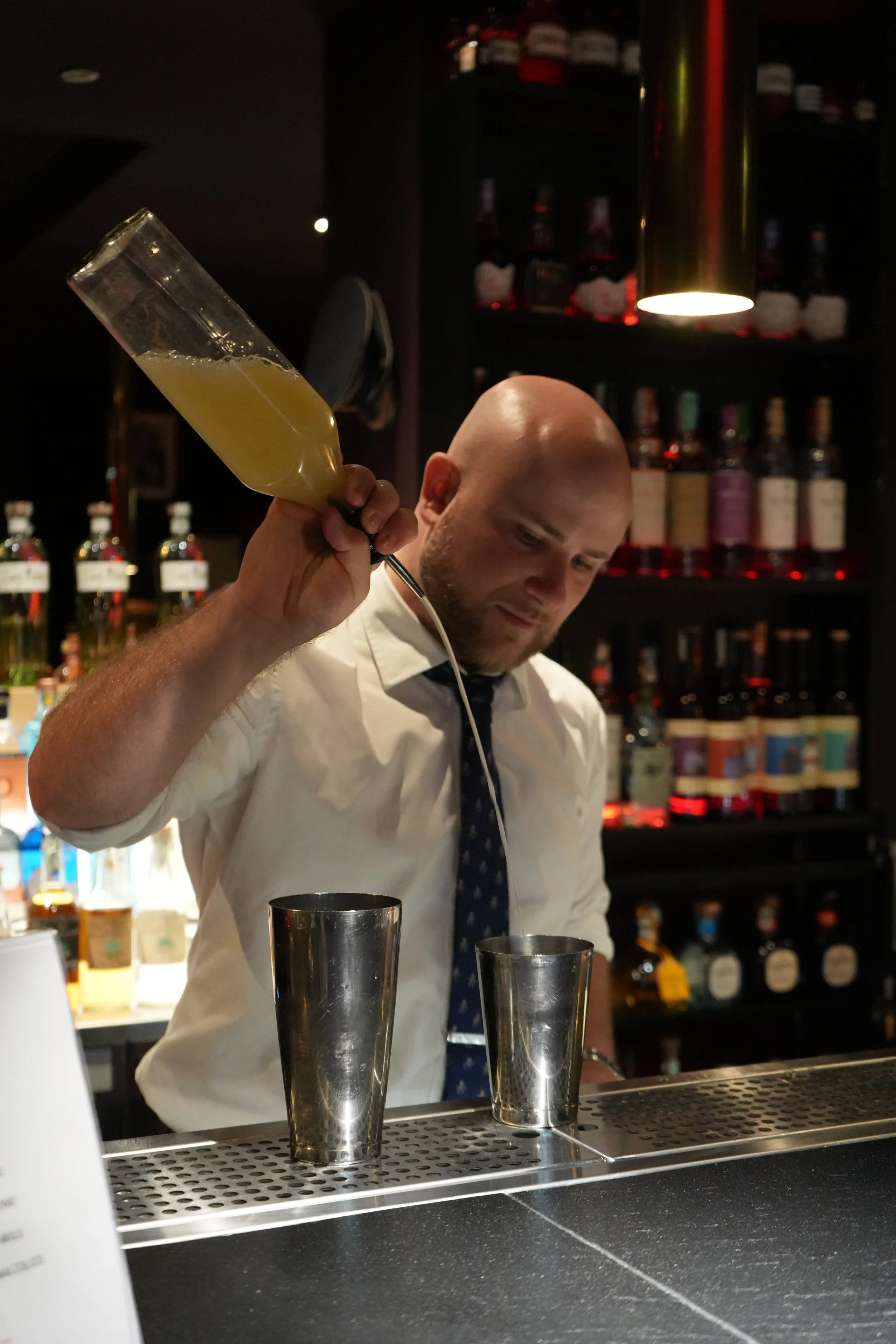 Bald bartender in white shirt and tie preparing drinks behind bar, holding a cocktail shaker in his right hand, with dark shelves of liquor bottles in the background, and two metal shakers on the bar counter.