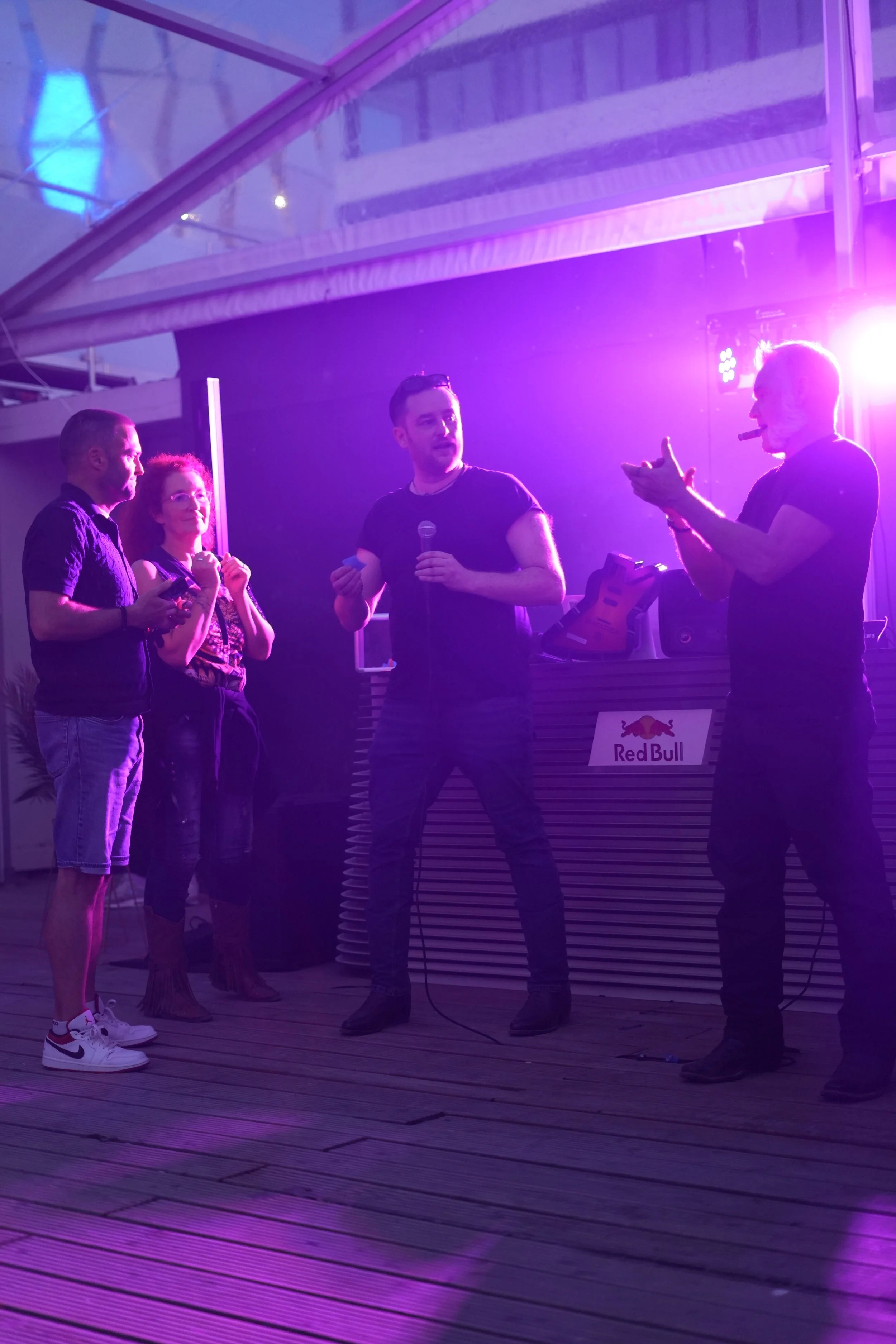 Four people standing in a semi-circle under purple lighting with a DJ booth and Red Bull logo in the background. One person appears to be speaking while the others listen and take photos or videos.