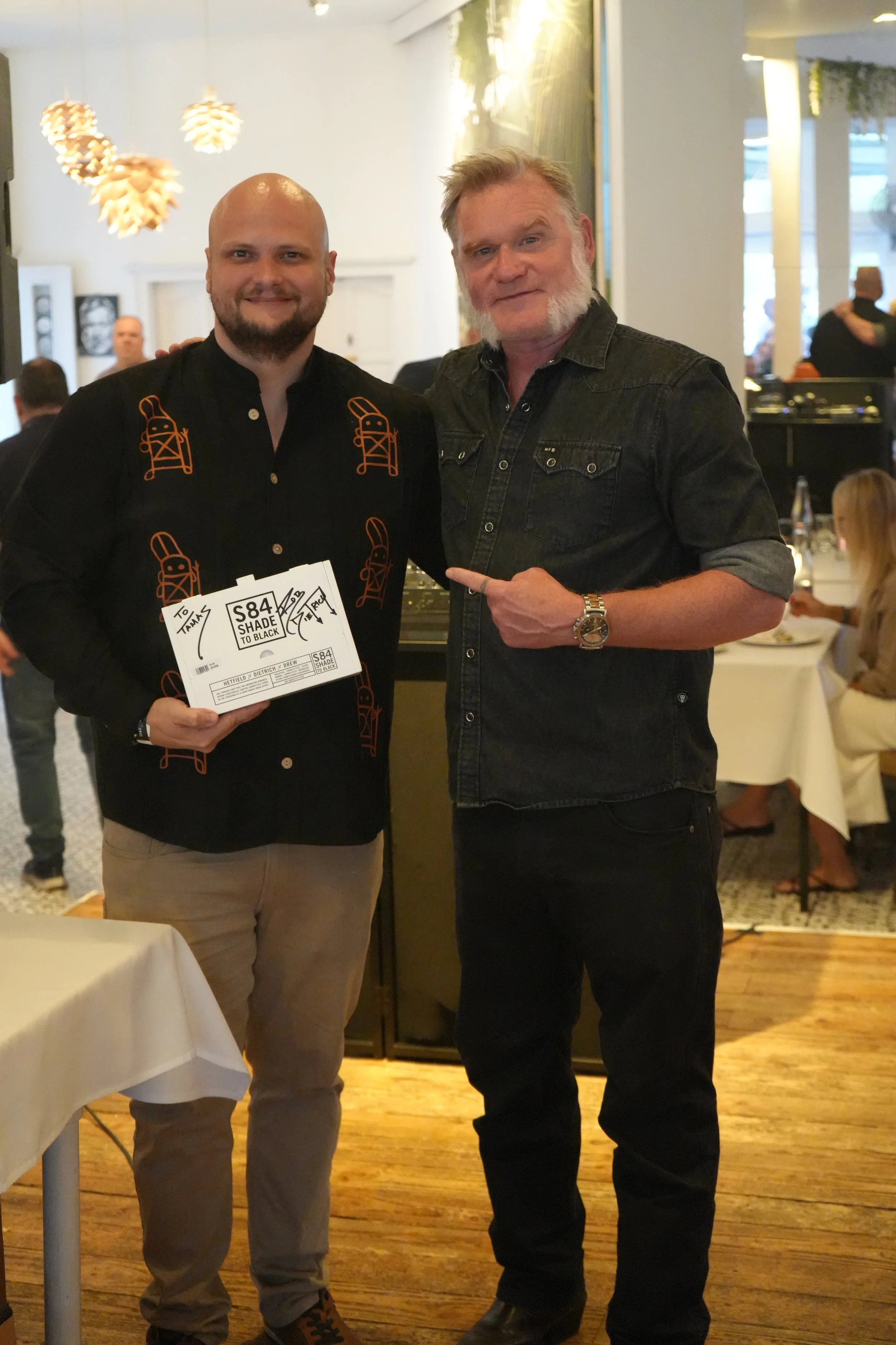 Two men standing together in a restaurant or event space. The man on the left is holding a ticket and is wearing a black shirt with orange designs. The man on the right has gray hair and beard, wearing a dark denim shirt with rolled-up sleeves, and p