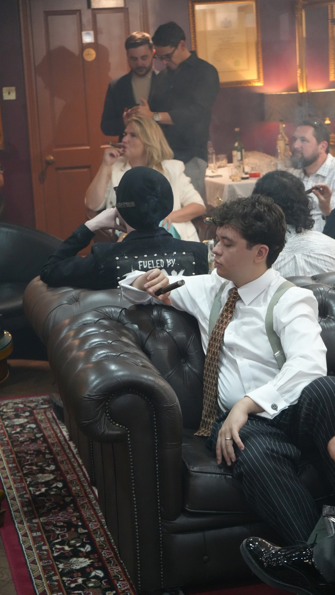 People in a dimly lit lounge, some sitting on a brown leather couch with cigarettes, one woman in white sitting at a table, some men standing in the background, and framed certificates on the wall.