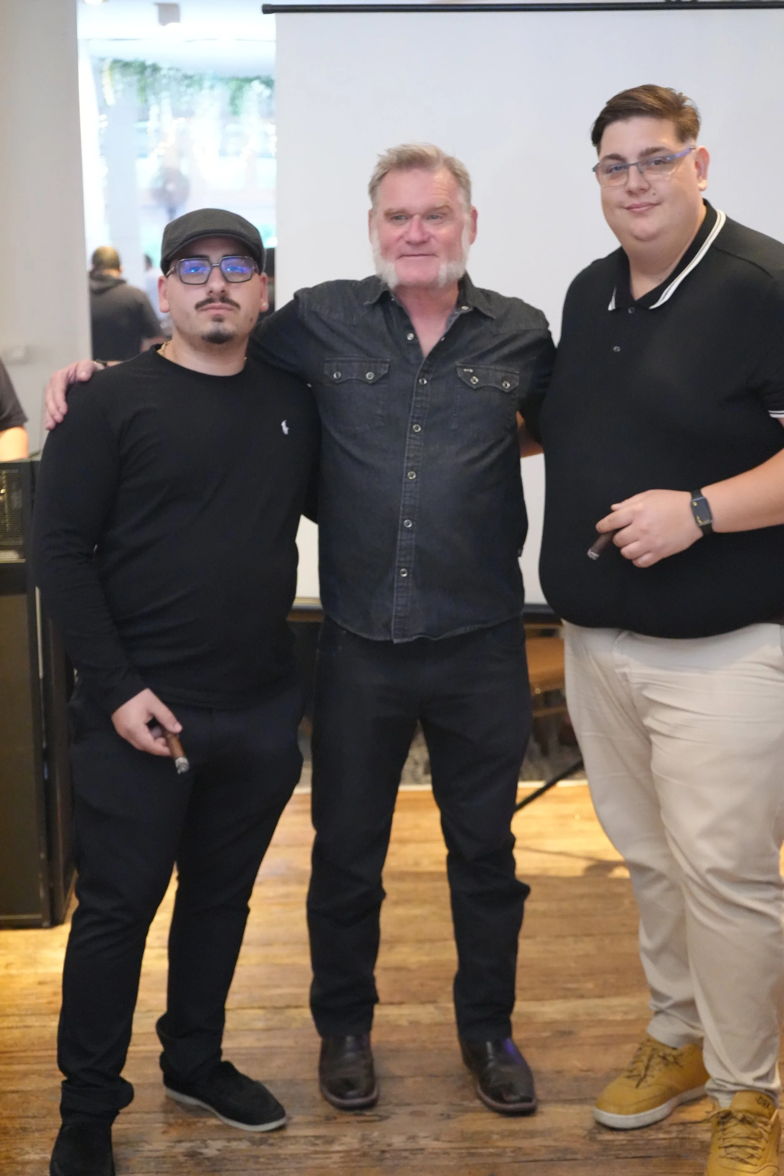 Three men standing together indoors, with one man wearing glasses and a black shirt, the second man in the middle with a gray beard and black shirt, and the third man wearing glasses and a black polo shirt with white trim, all posing for a photo.