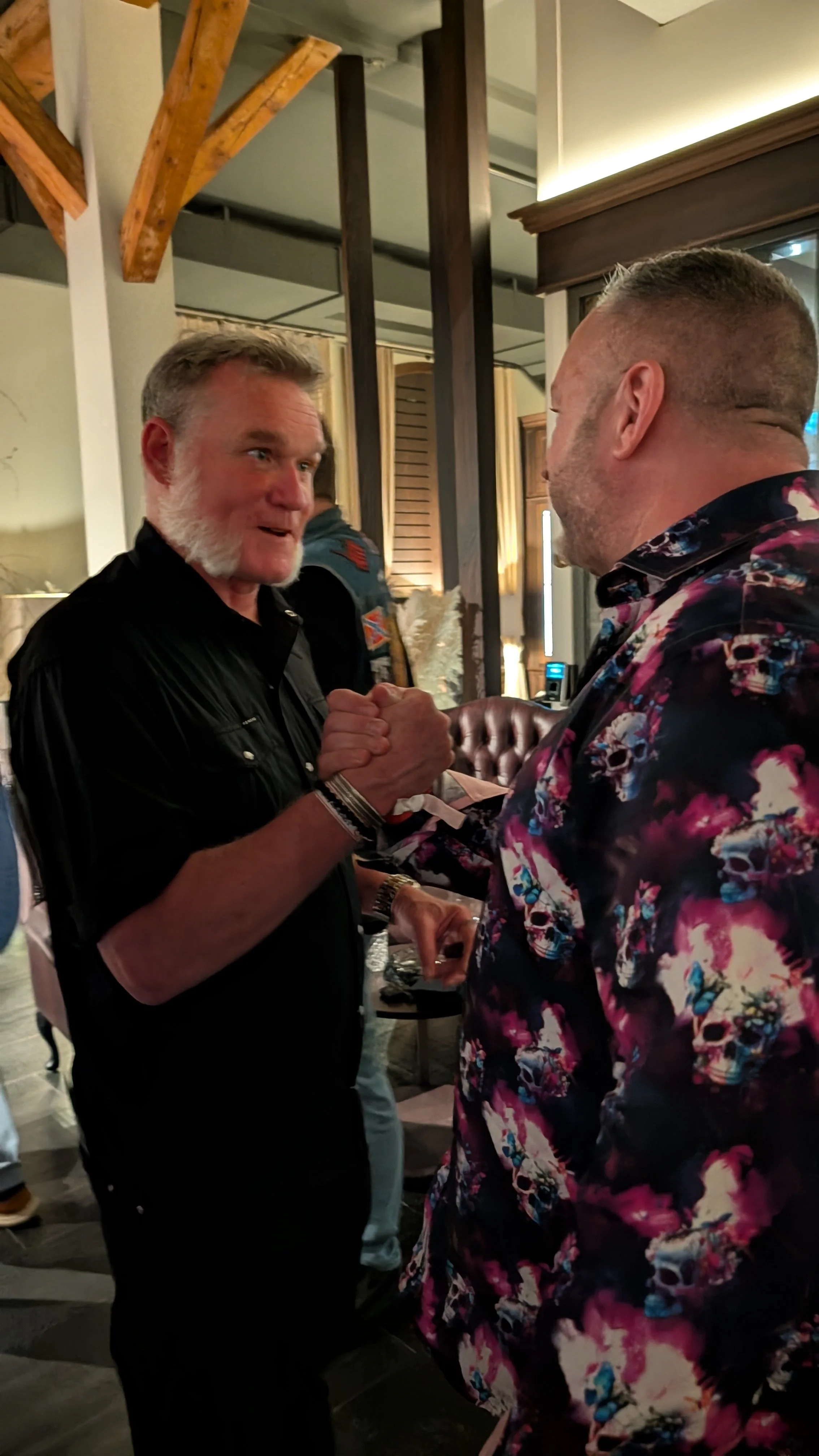 Two men are shaking hands and smiling in a social setting, possibly a restaurant or bar, with warm lighting and wooden decor.