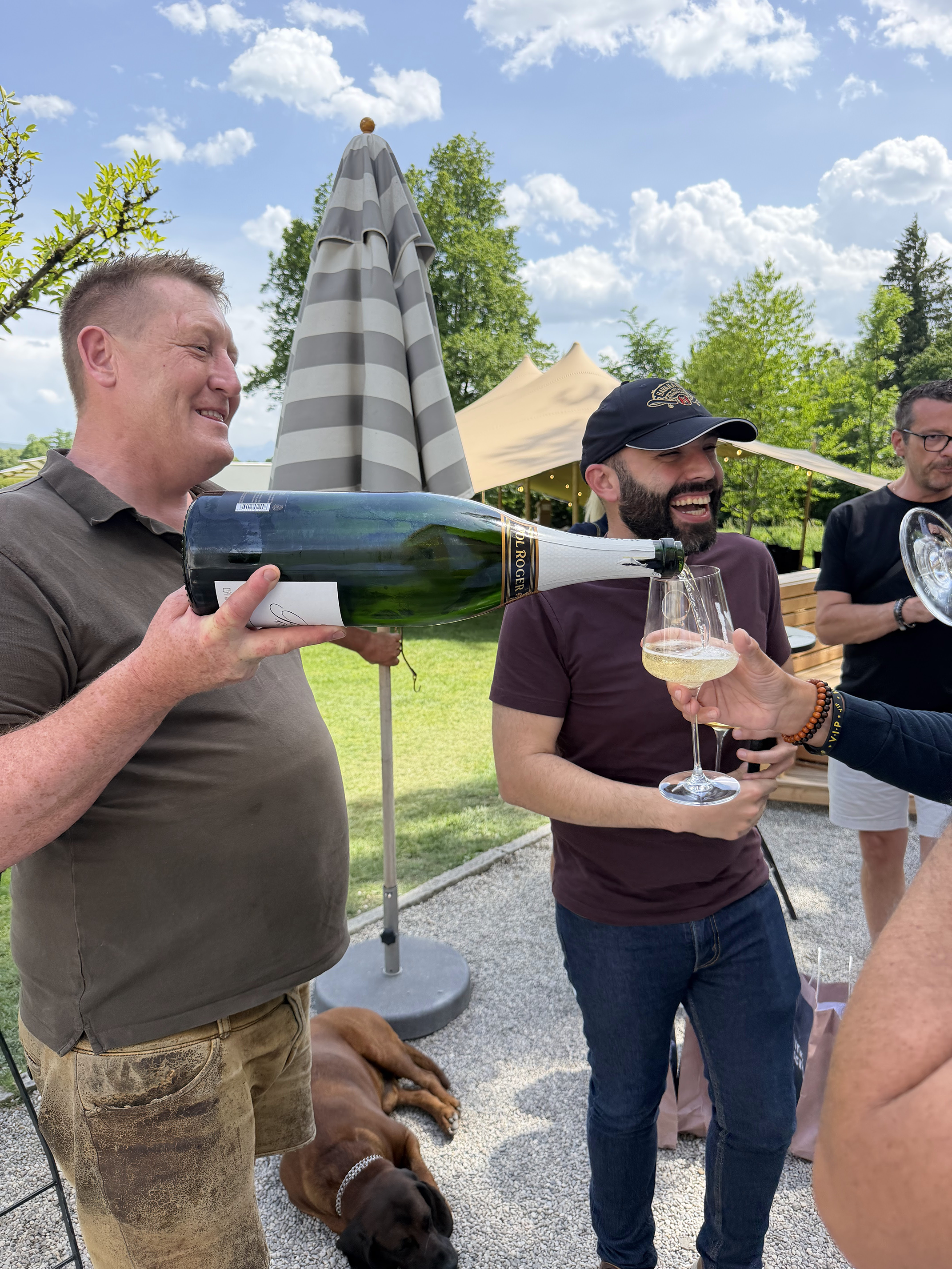 Men at an outdoor gathering pouring and enjoying wine, with dogs resting on the ground nearby, surrounded by trees and tents.