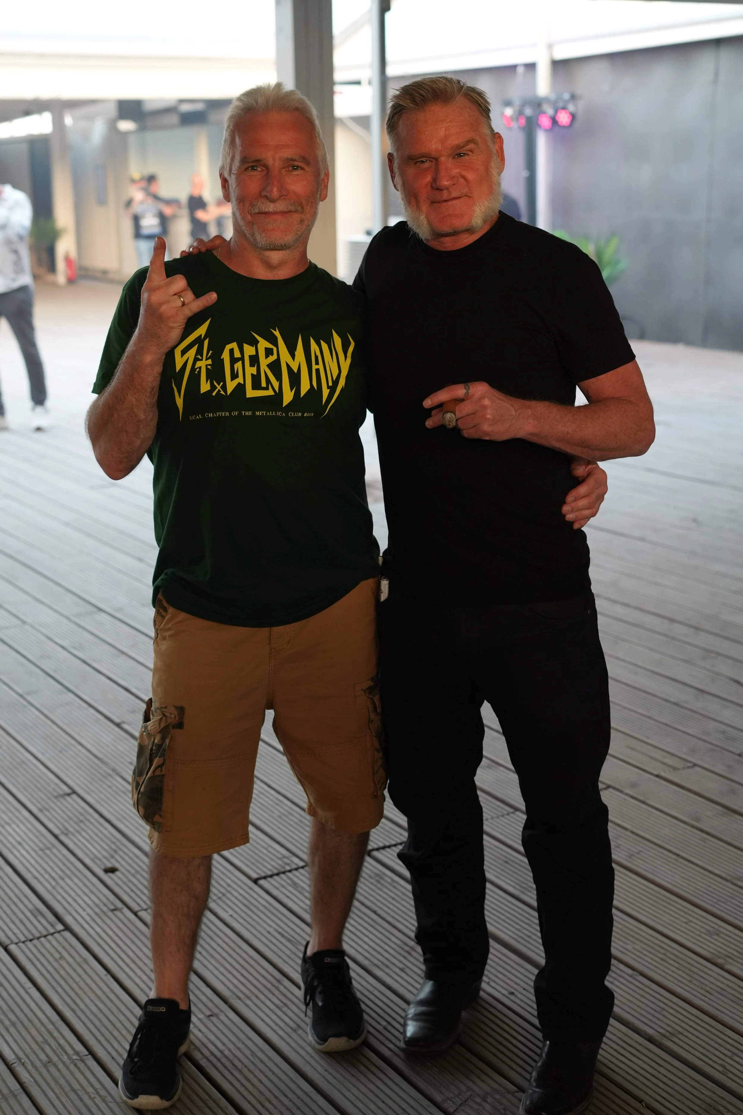 Two older men with gray hair and beards standing close together, smiling, at an indoor or covered outdoor event, with one wearing a green Metallica T-shirt and the other in a black shirt, both making gestures with their hands.