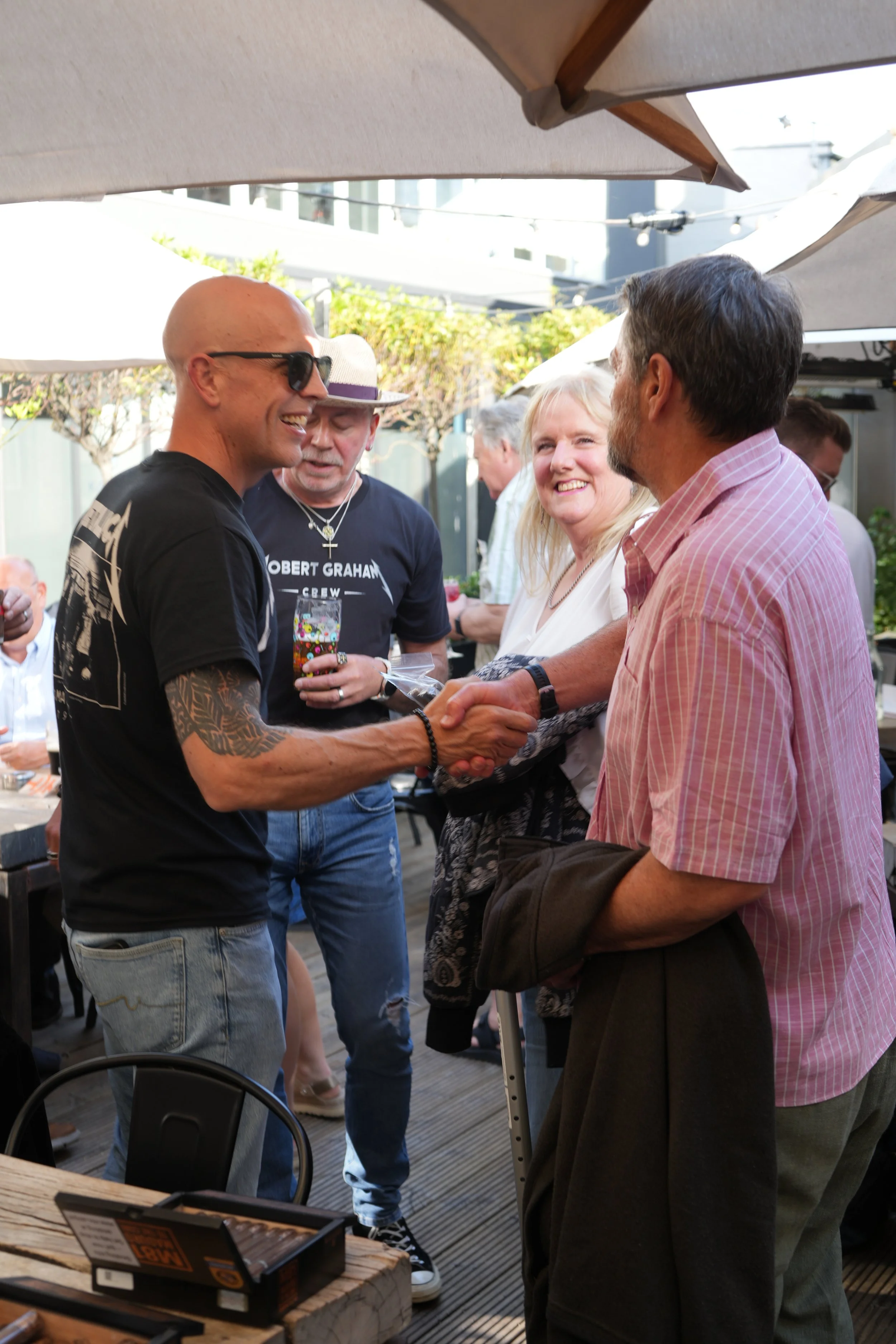 A group of people socializing outdoors under umbrellas, shaking hands, smiling, and holding drinks at a gathering or party.
