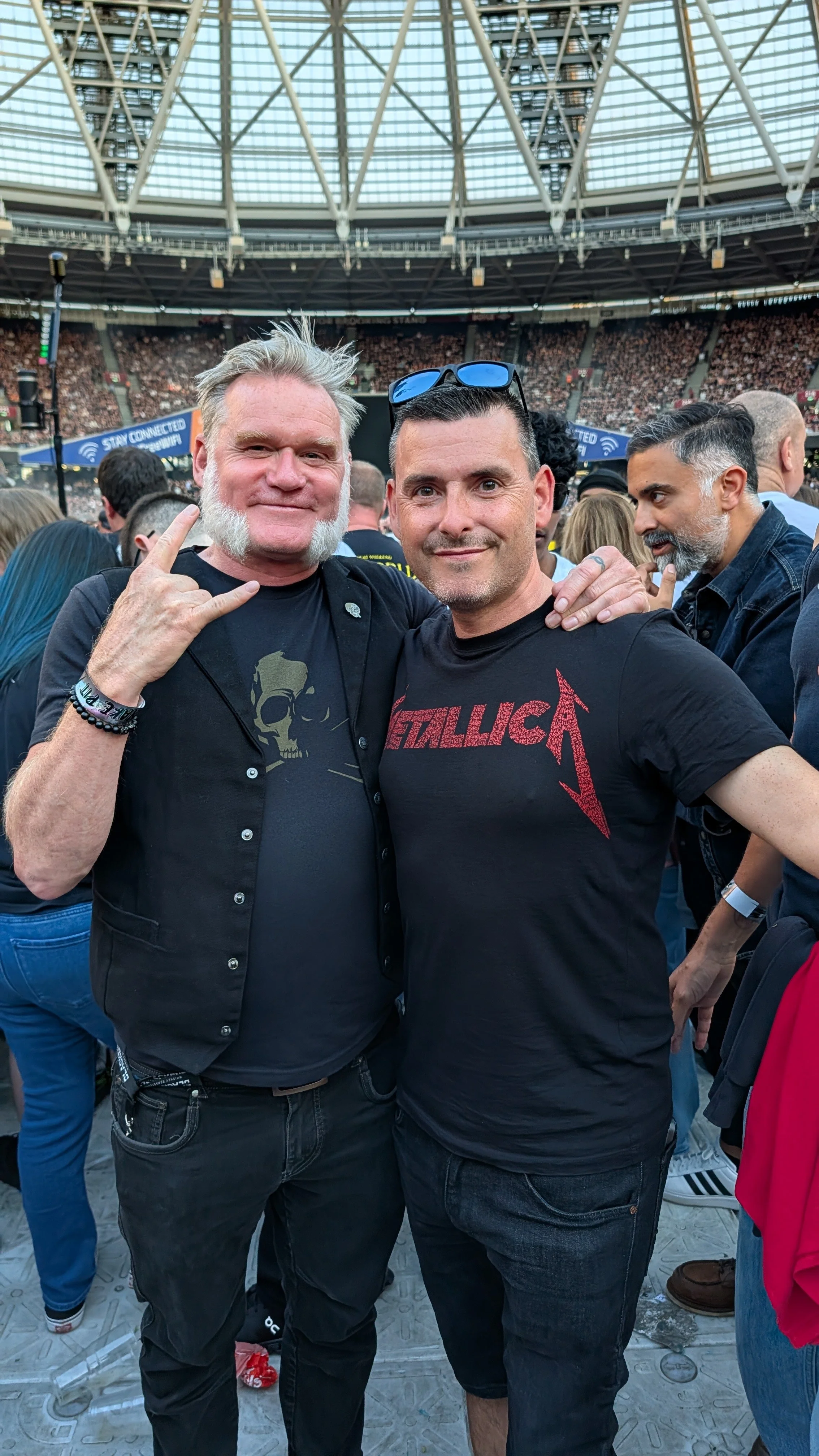 Two men standing together at a crowded concert venue, smiling at the camera. One is making a rock gesture with his hand, wearing a black shirt with a skull graphic. The other is wearing a black Metallica T-shirt. The background shows a large crowd, s