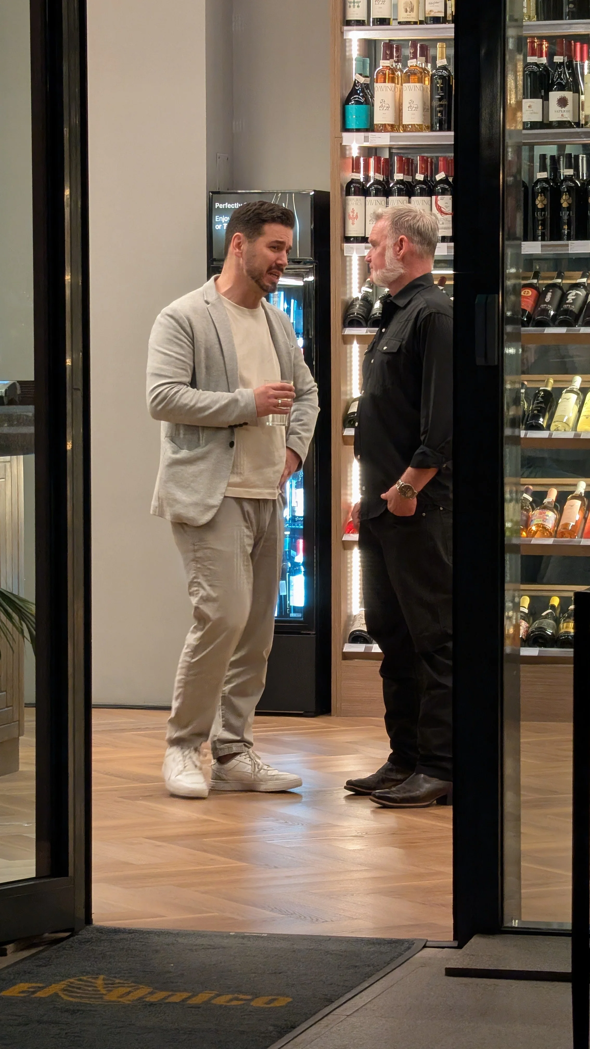 Two men are standing and talking inside a wine shop, with wine bottles on shelves behind them.