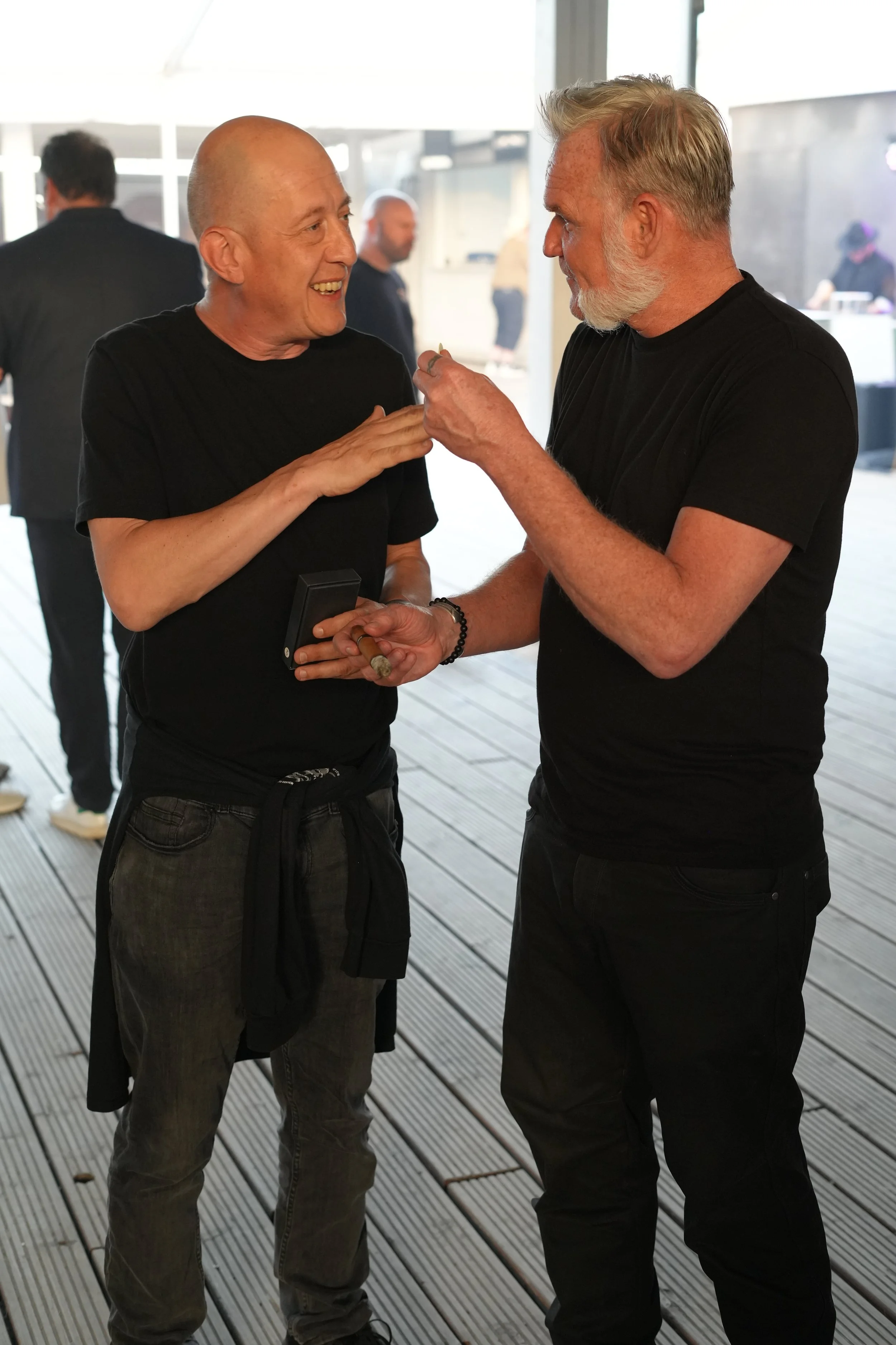 Two middle-aged men having a conversation, smiling and laughing at an indoor event or gathering, with other blurred people in the background.