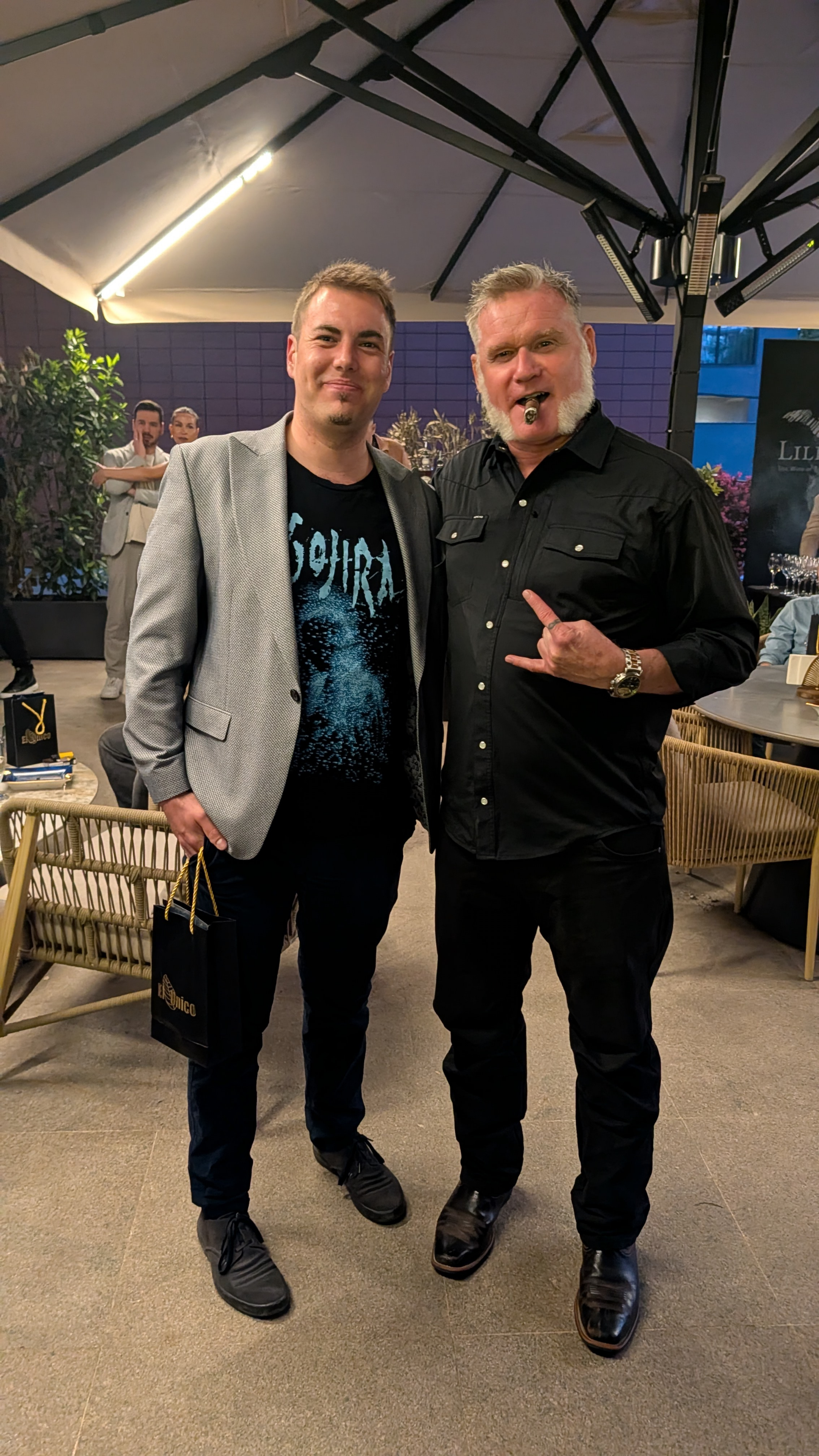 Two men standing together at an indoor event, one wearing a light gray blazer and black t-shirt, the other in a black button-up shirt with a cigar in his mouth, and both making a hand gesture.