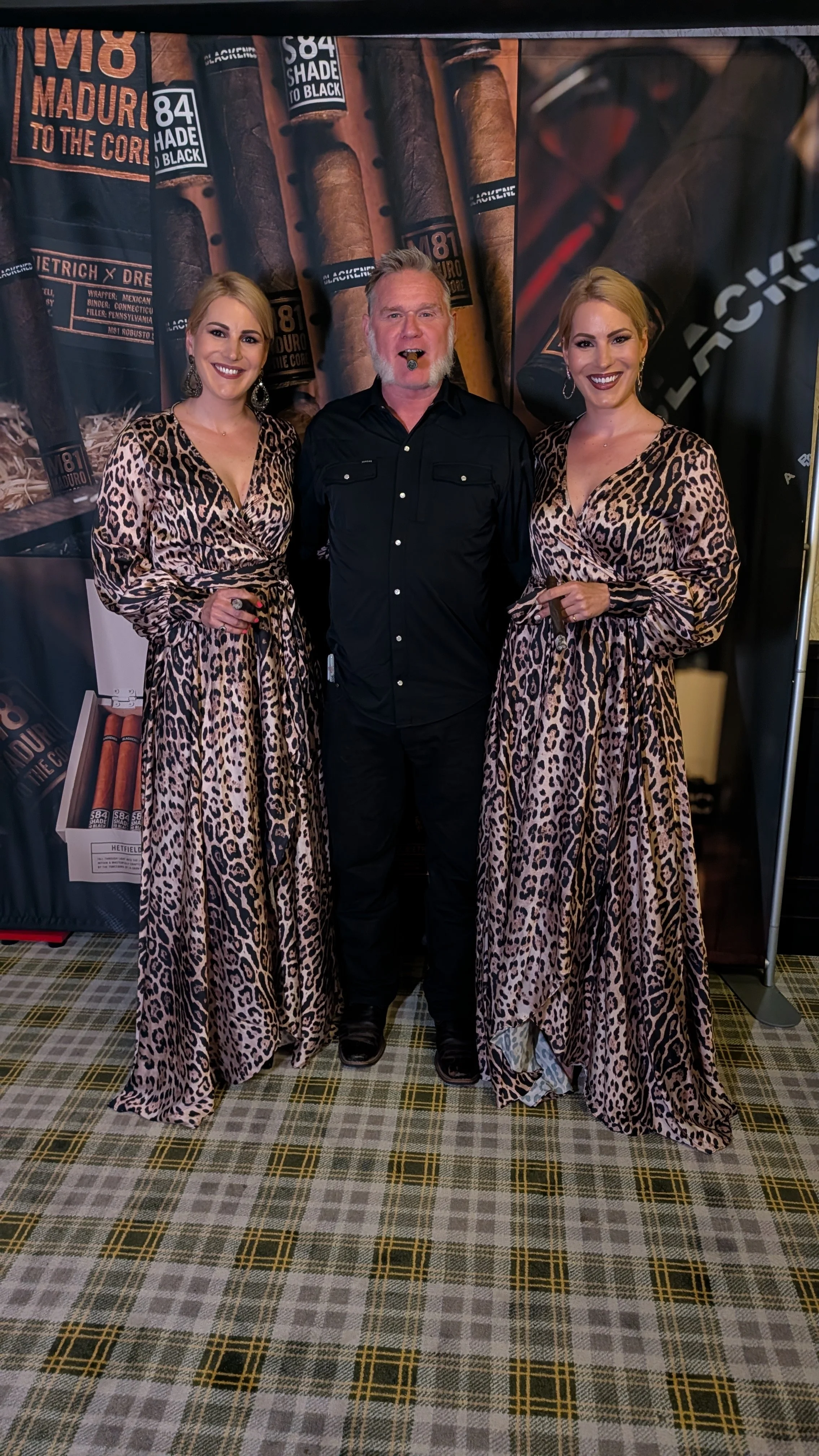 Three people, two women and one man, standing in front of a backdrop with cigar images and labels. The women are wearing matching leopard print dresses, and the man is dressed in black, smoking a cigar.