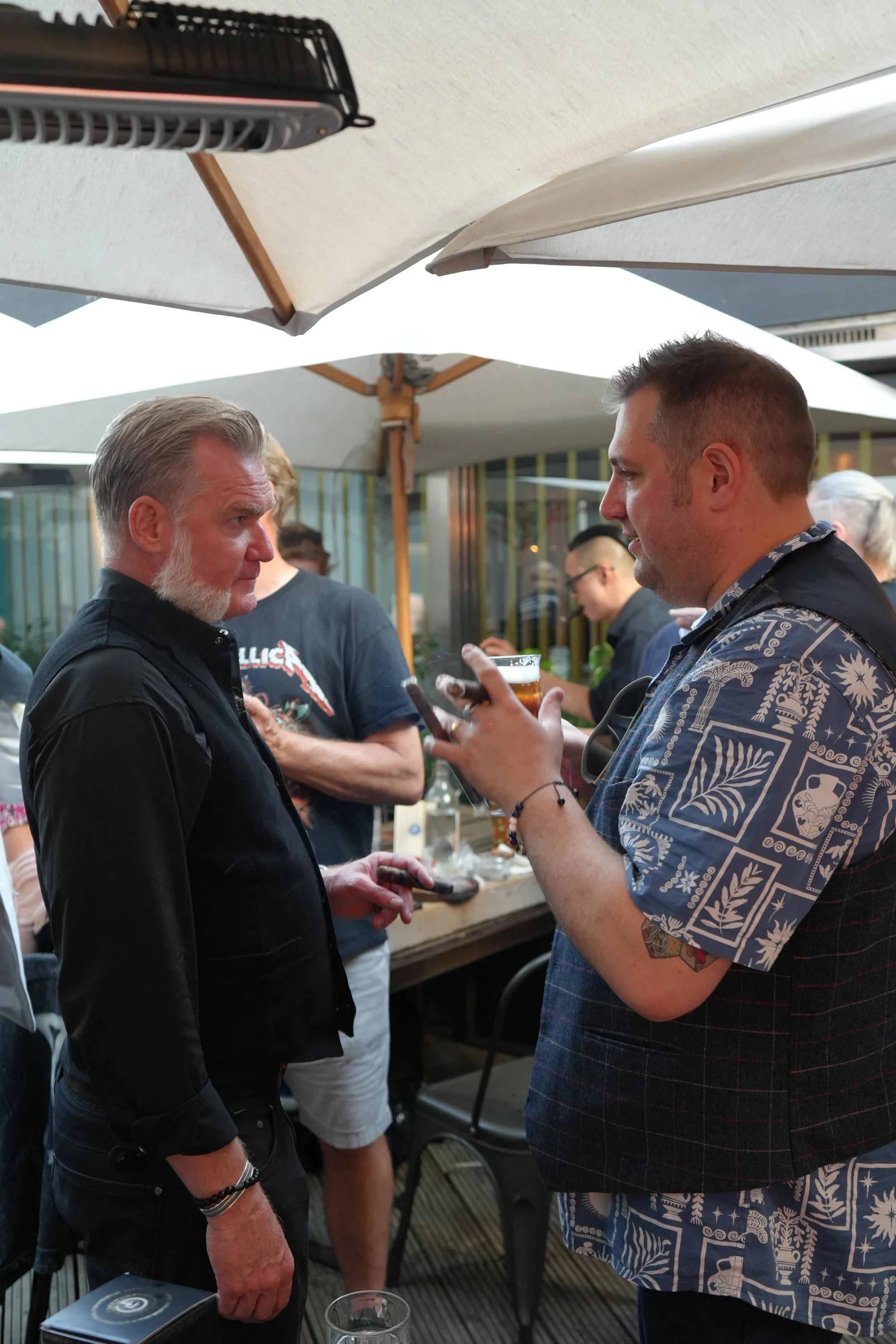 Two men having a conversation at an outdoor gathering, with two large umbrellas overhead, some other people in the background, and a bar or food stand behind them.