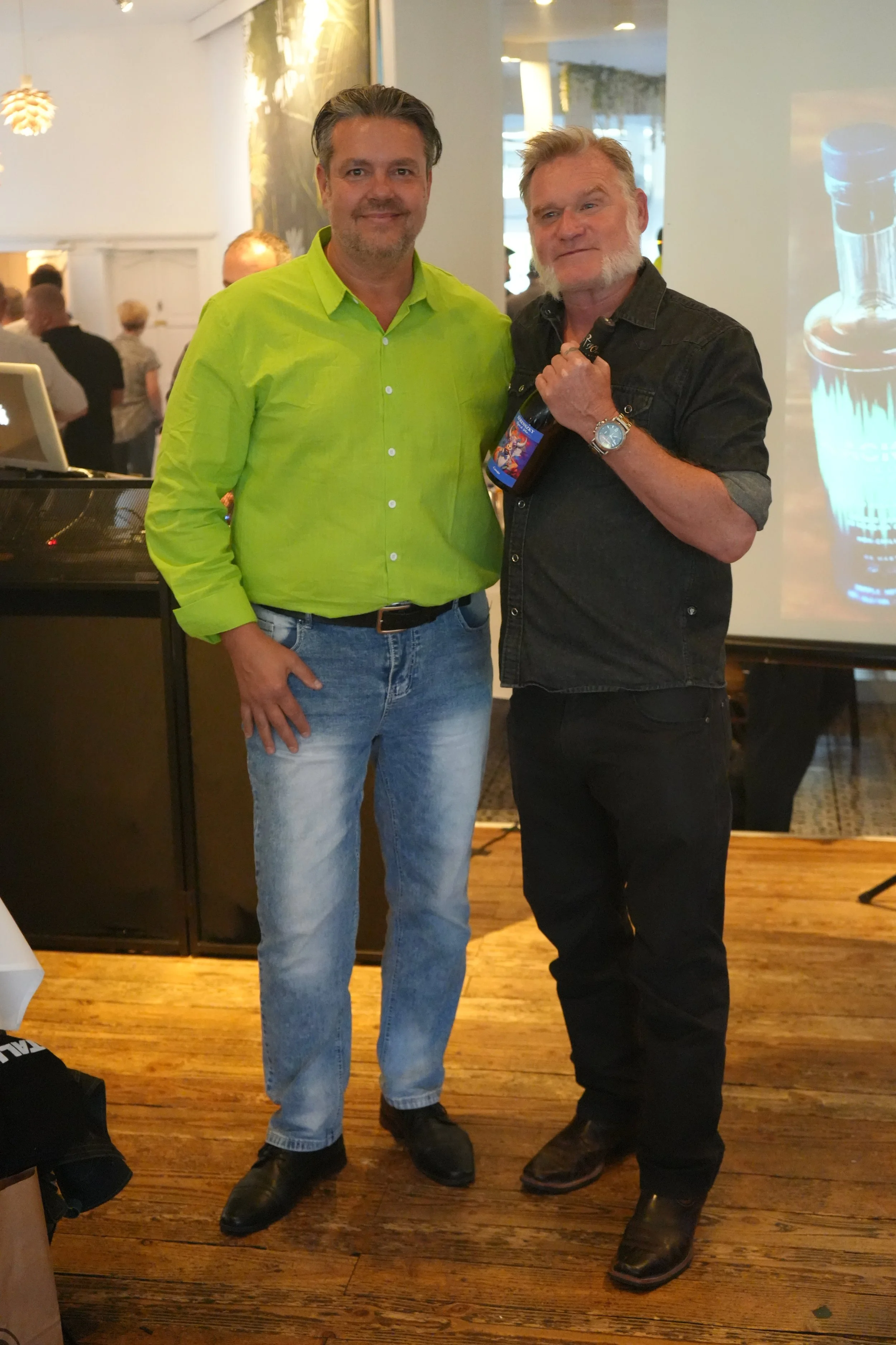 Two men standing inside a restaurant or event space, with one holding a bottle of beer, smiling at the camera. The man on the left wears a bright yellow-green shirt and blue jeans, while the man on the right wears a black denim shirt and black pants,