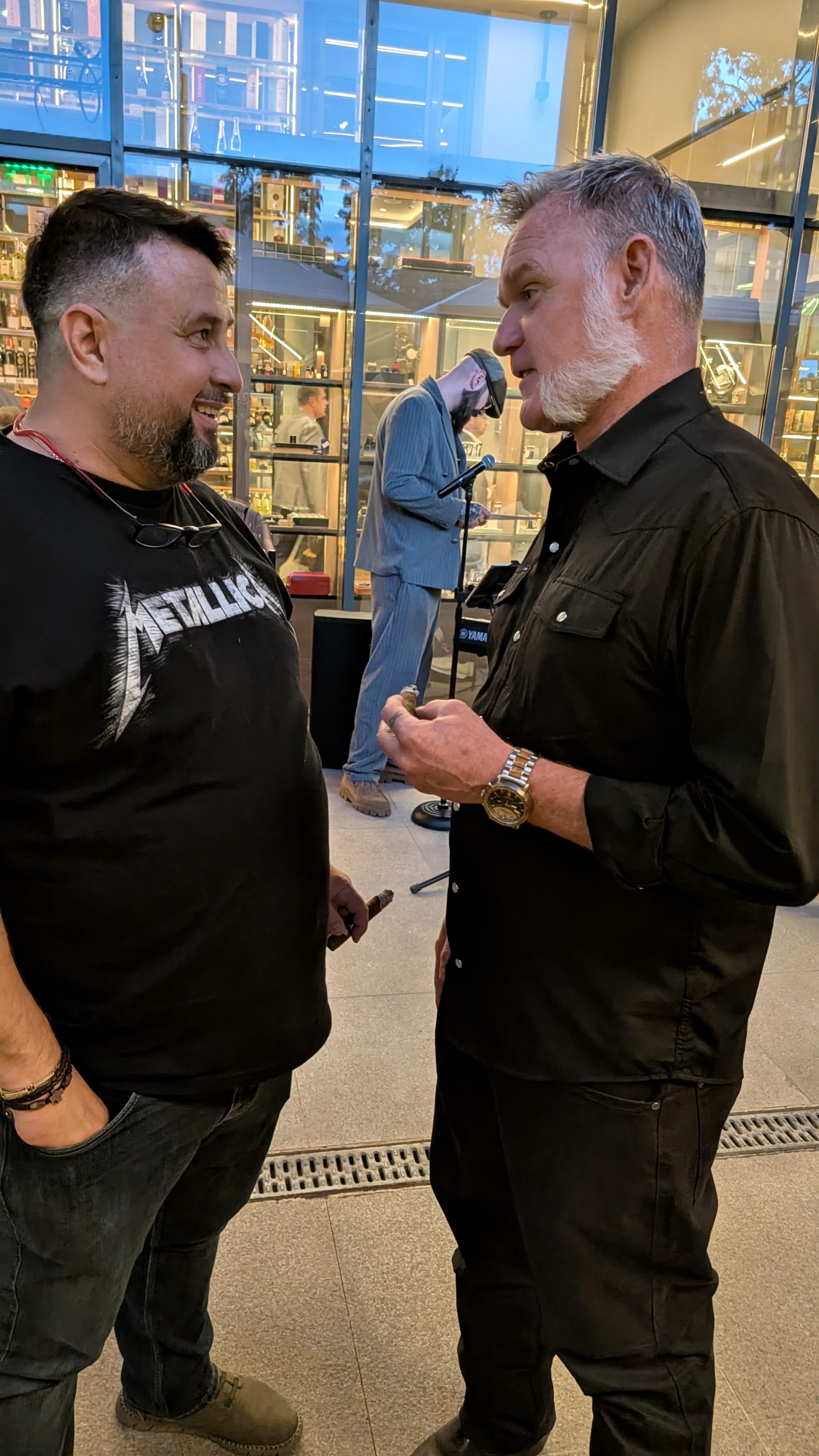 Two men engaged in conversation indoors with a glass wall in the background showing a store or cafe interior and a man using a microphone in the background.